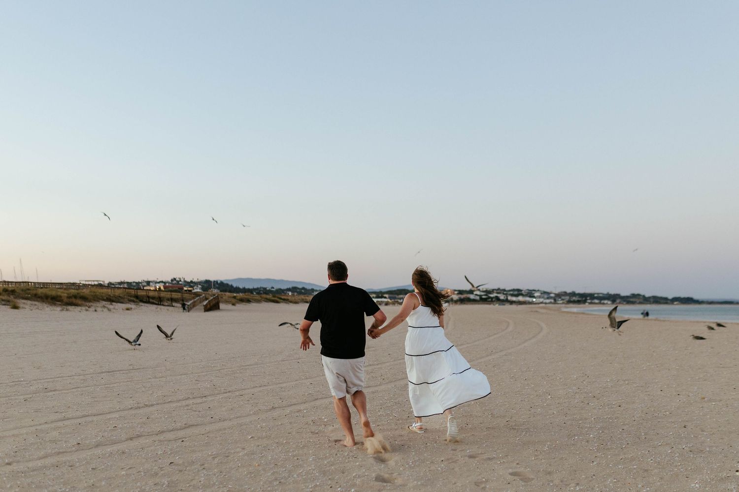 Couple chasing each other on beach with seabirds flying in background at dusk. Meia Praia, Lagos, Algarve, Portugal.