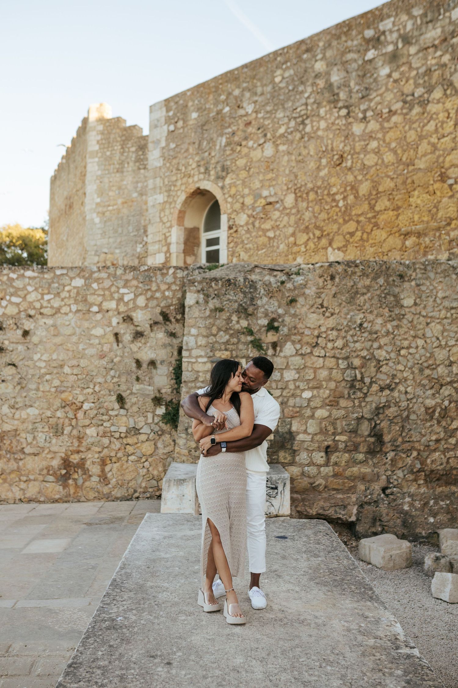 A romantic couple in white outfits embraces while standing against ancient stone fortress walls at sunset.