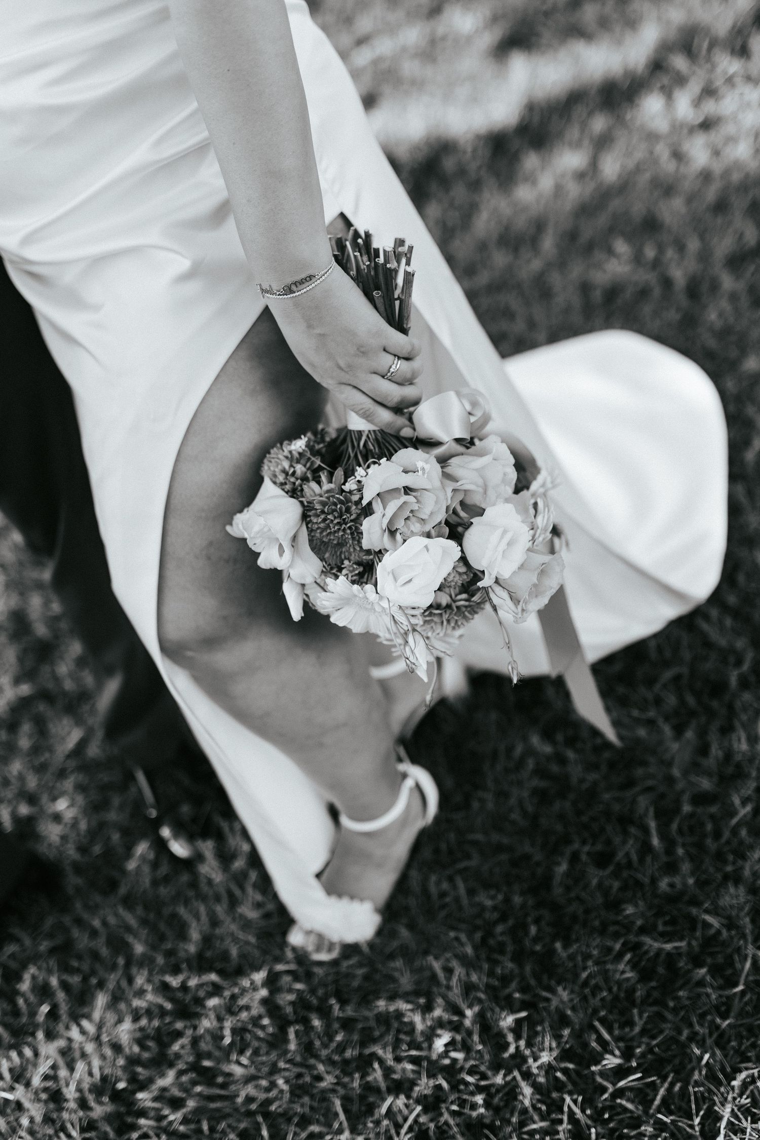 A close-up black and white shot of a bride holding a bouquet of roses and wearing white heels on grass.