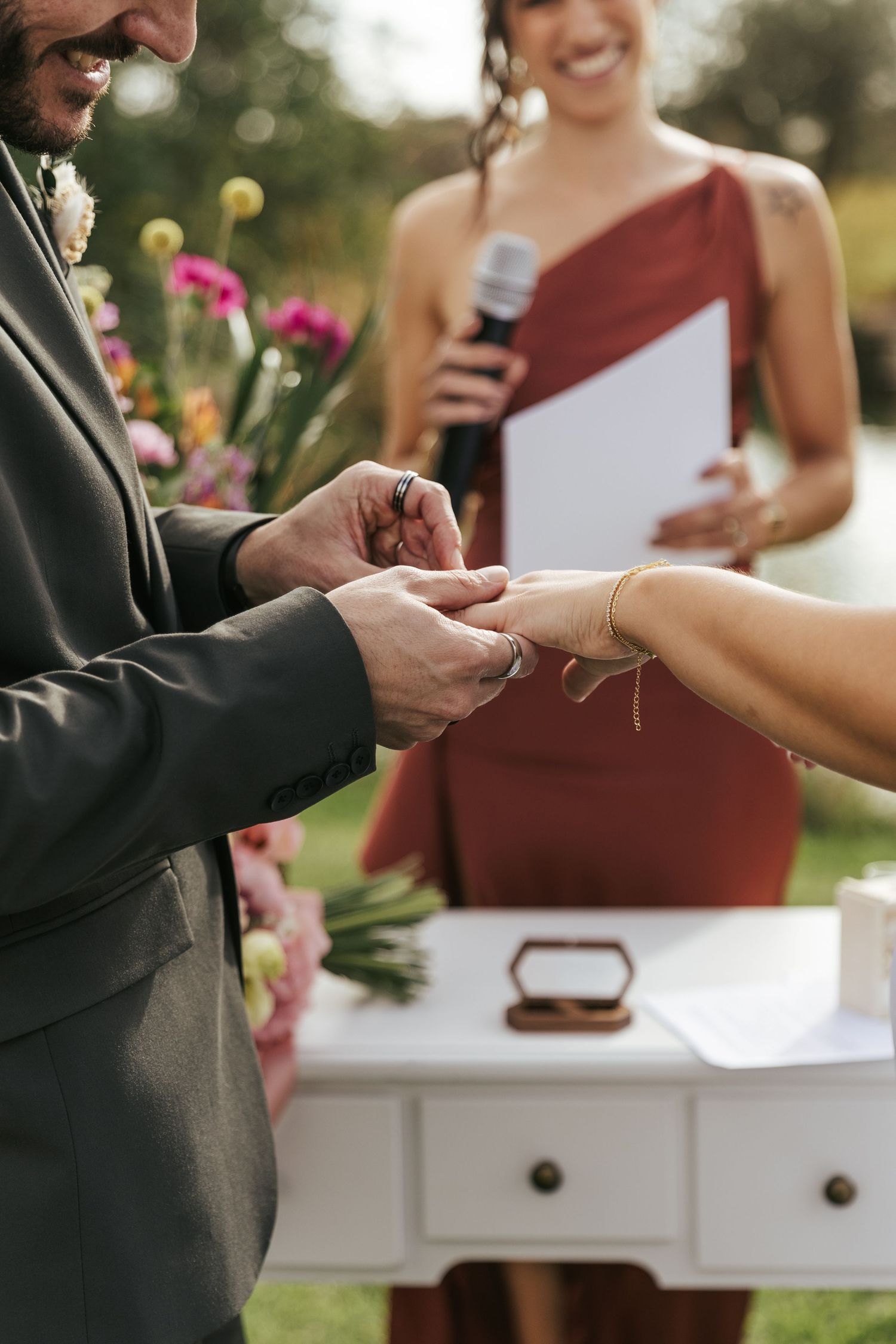 Close up of a ring exchange during an outdoor wedding ceremony with flowers in the background.