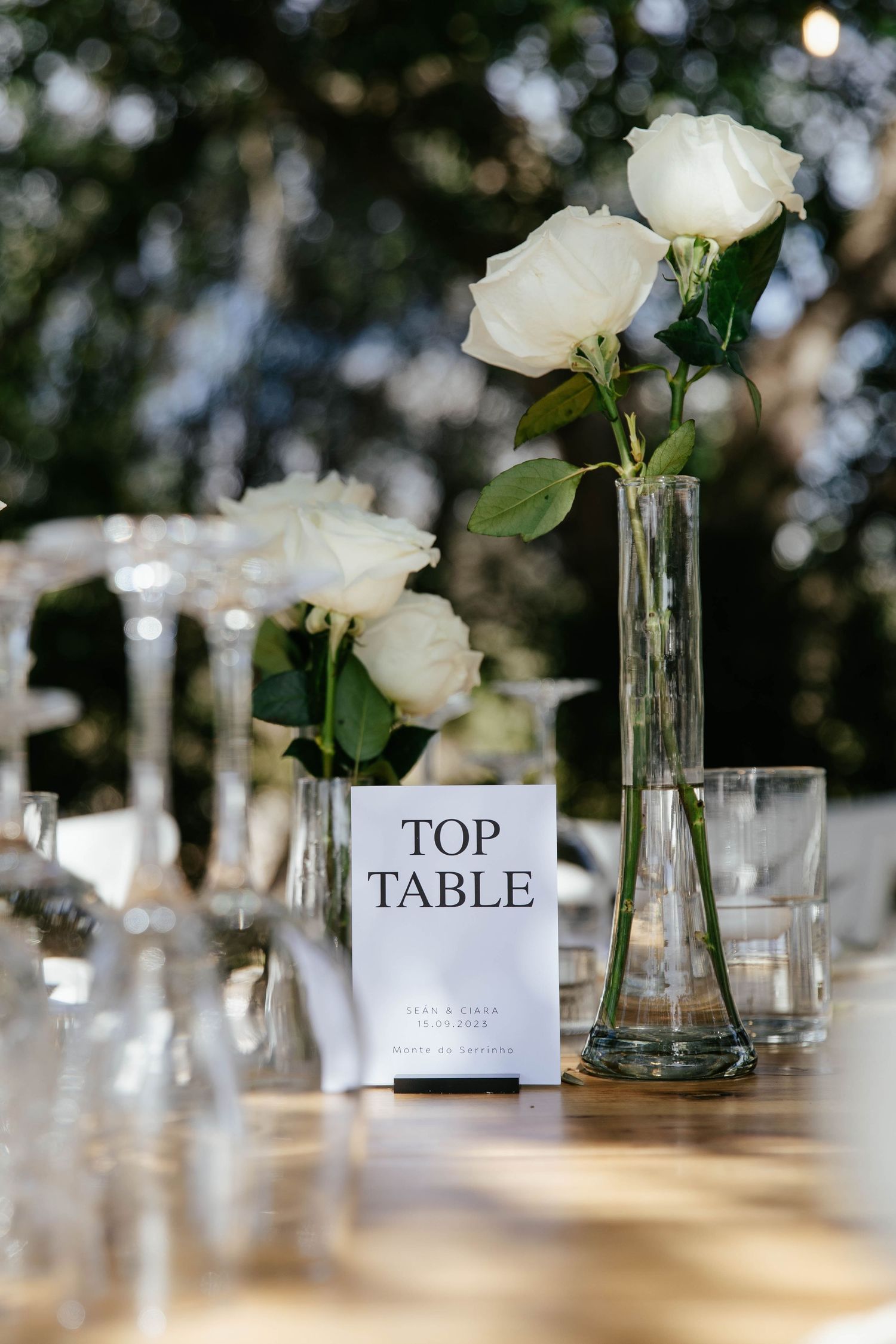 White roses in glass vases line a wedding reception table with a 'Top Table' sign.