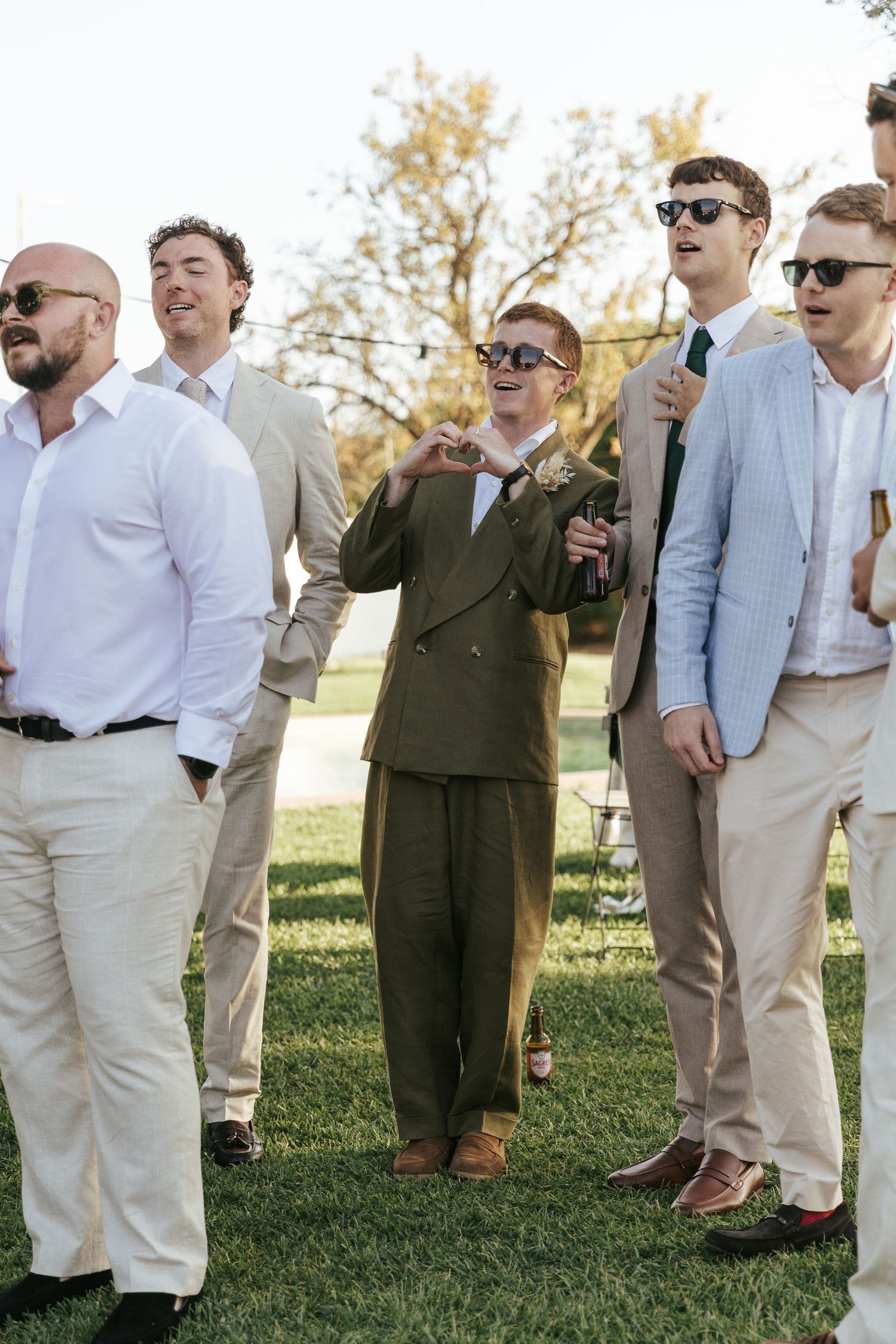 Group of men in suits celebrating outdoors at a wedding, one making a heart gesture.