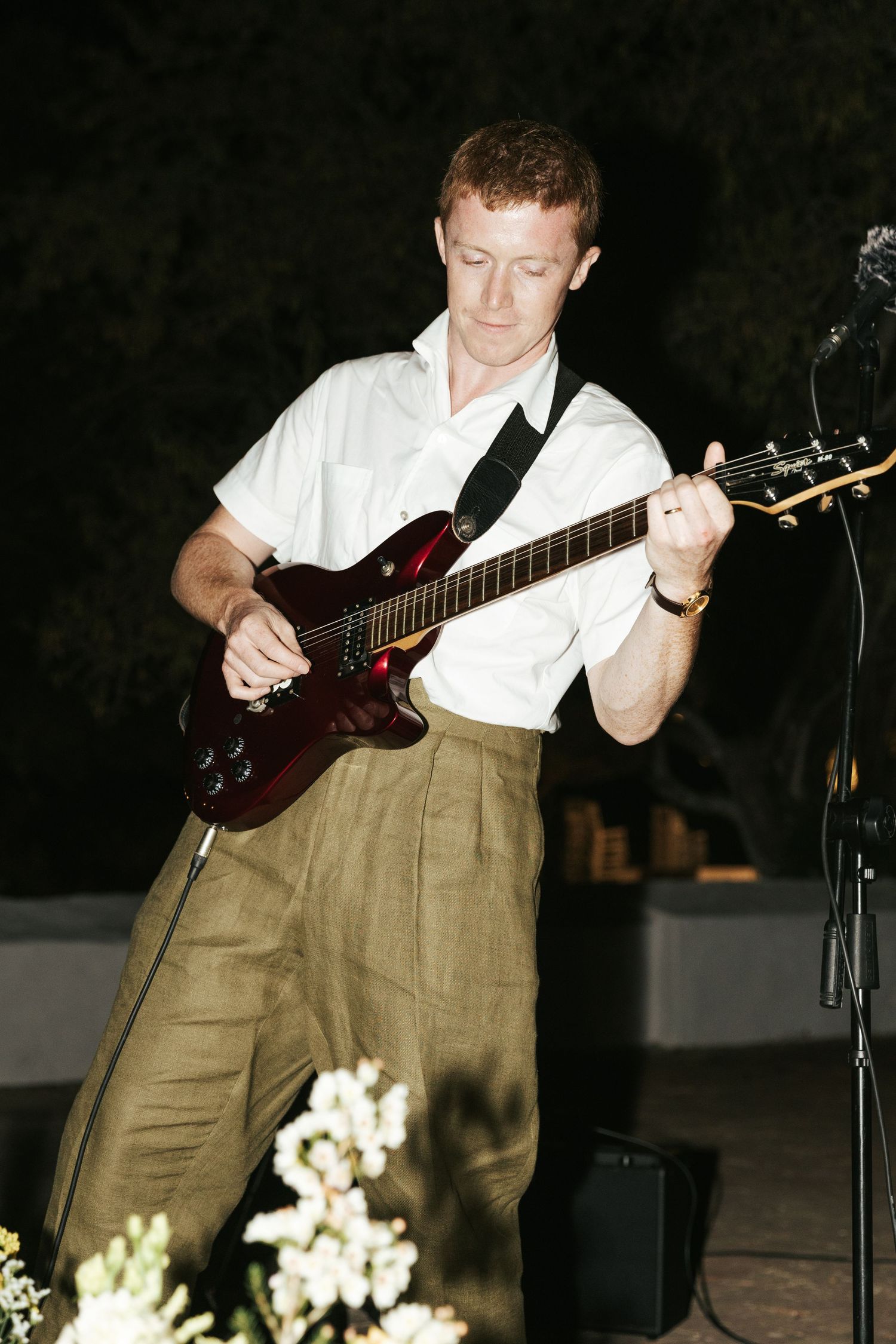 A young man in a white shirt and khaki pants plays an electric guitar at an outdoor evening event.