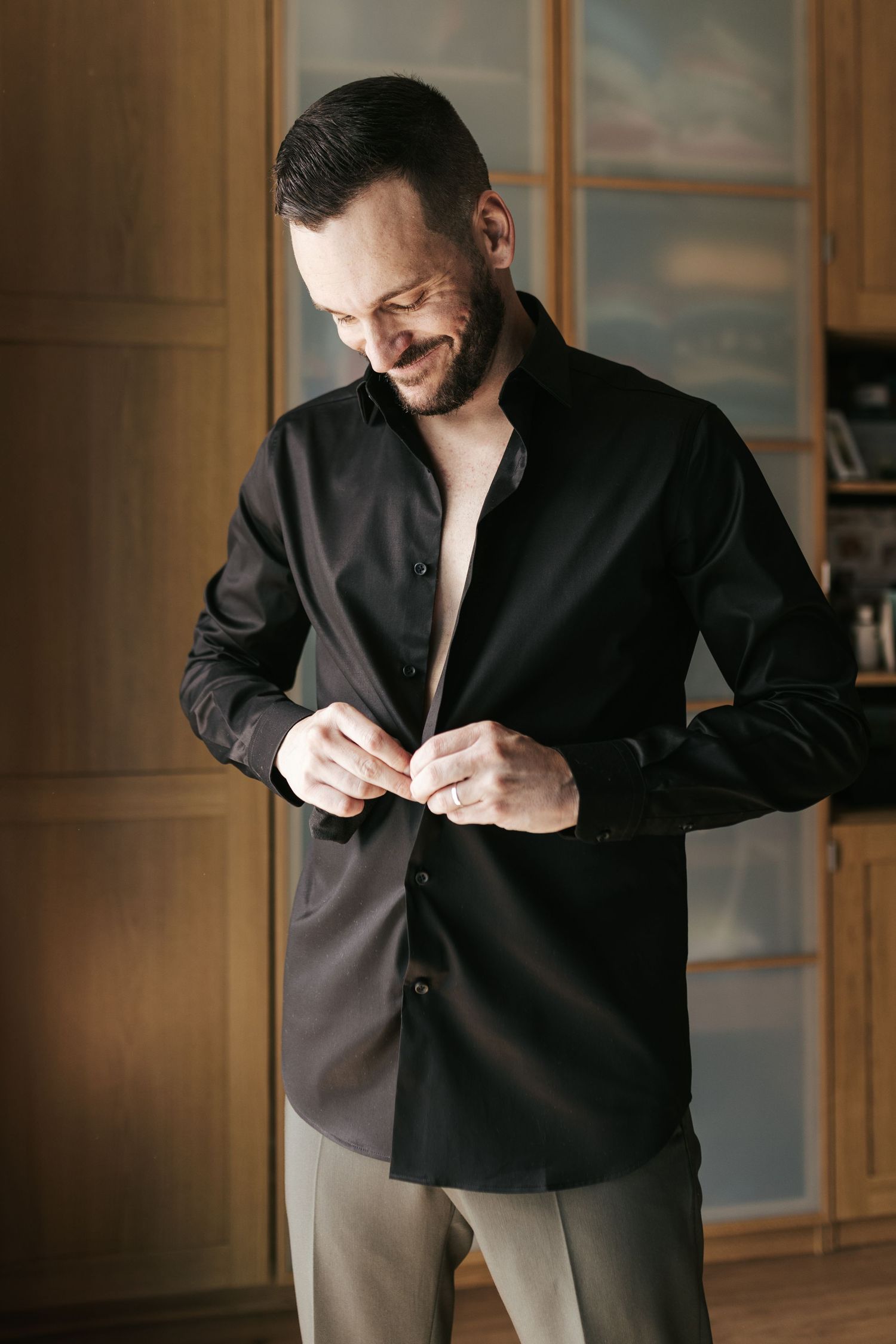 Smiling person in black button-up shirt buttons their clothing while standing in front of wooden closet doors.