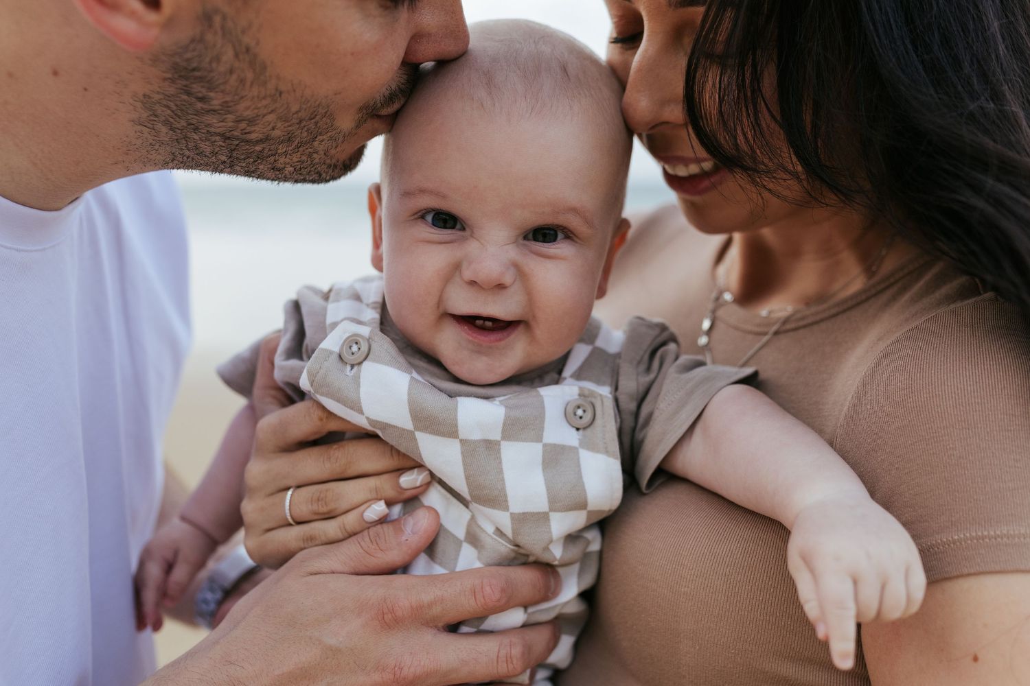 A smiling baby wearing a checkered outfit is held lovingly between two parents.