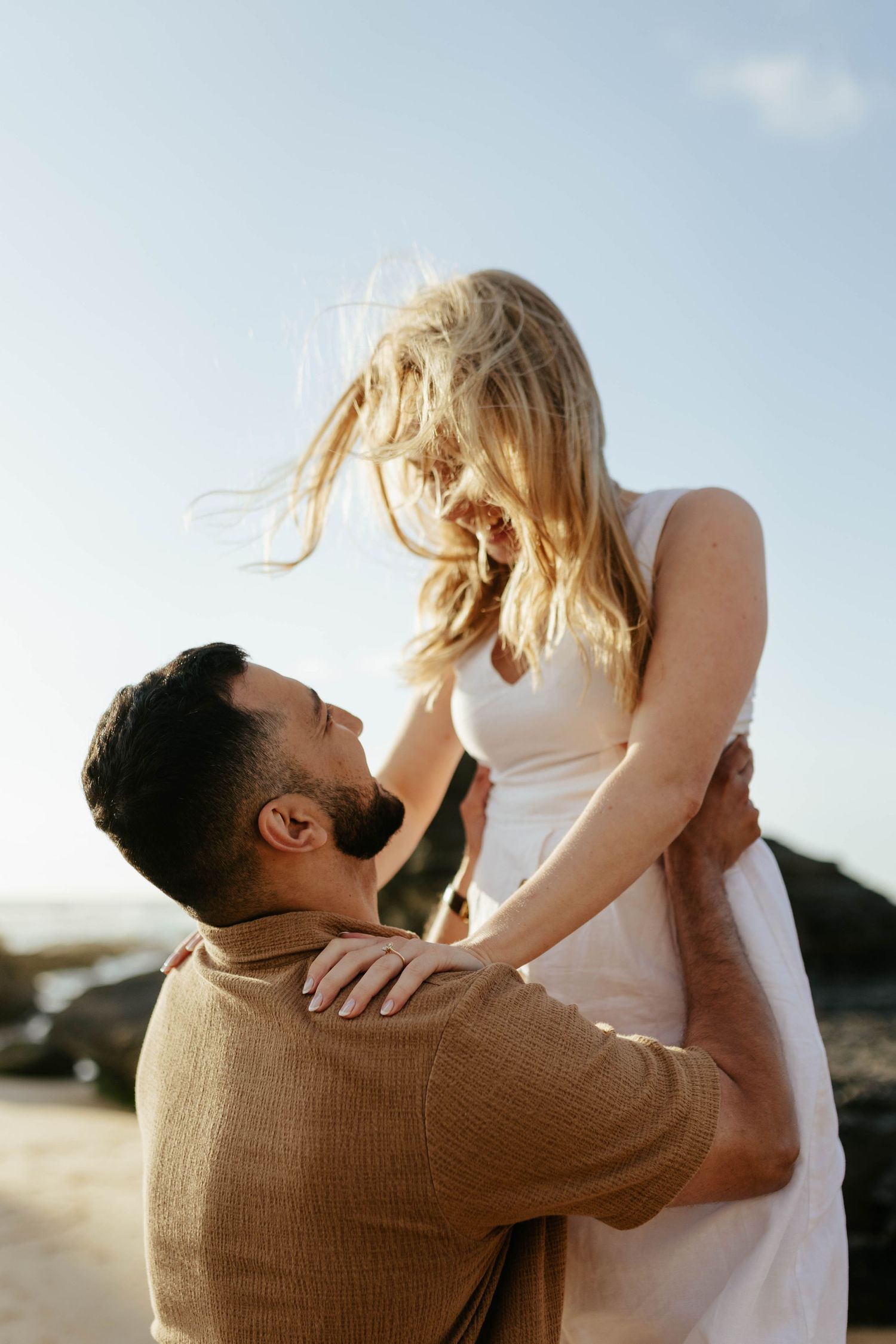 Romantic couple shares an intimate moment at sunset as he lifts her up in white dress on the beach.