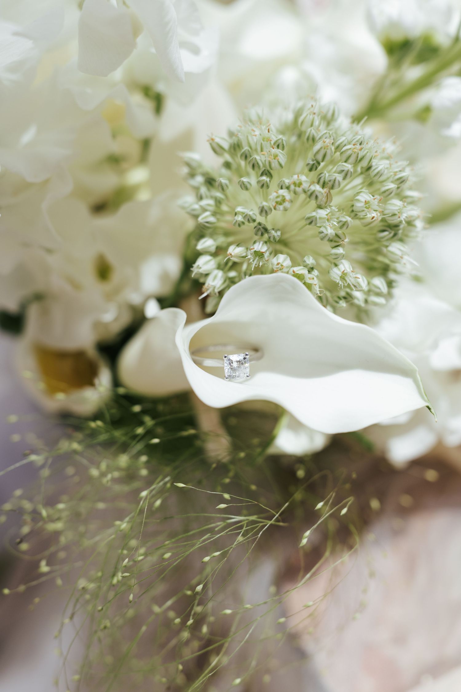 Diamond engagement ring resting in a white calla lily surrounded by elegant bridal bouquet of white flowers.