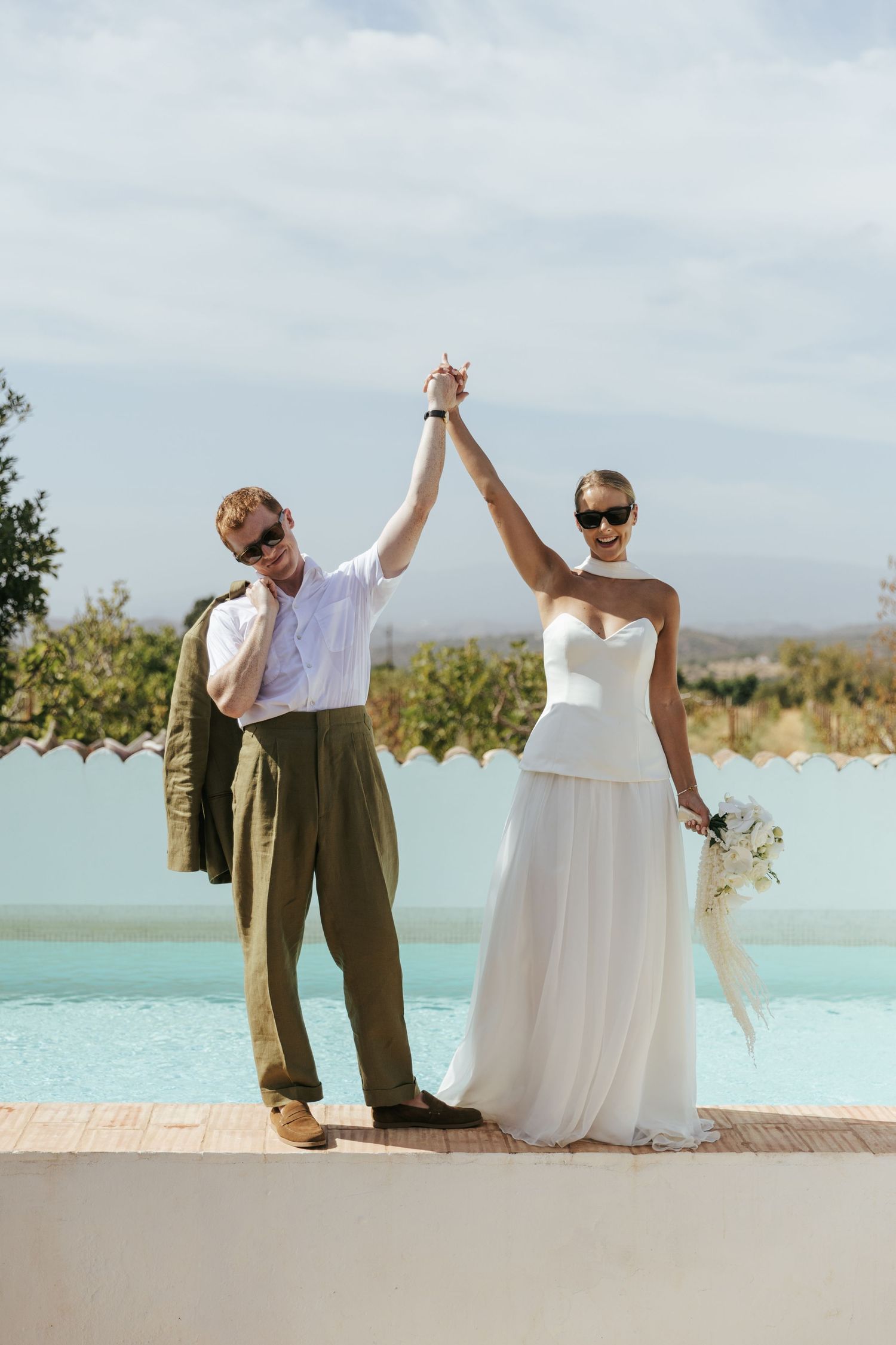 Bride and groom celebrate in sunglasses, raising hands by a pool at a sunny outdoor wedding venue.