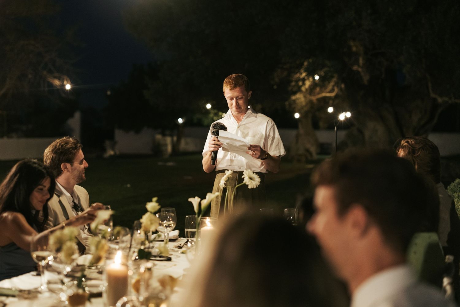Man delivering speech at candlelit outdoor wedding reception dinner under string lights at night.