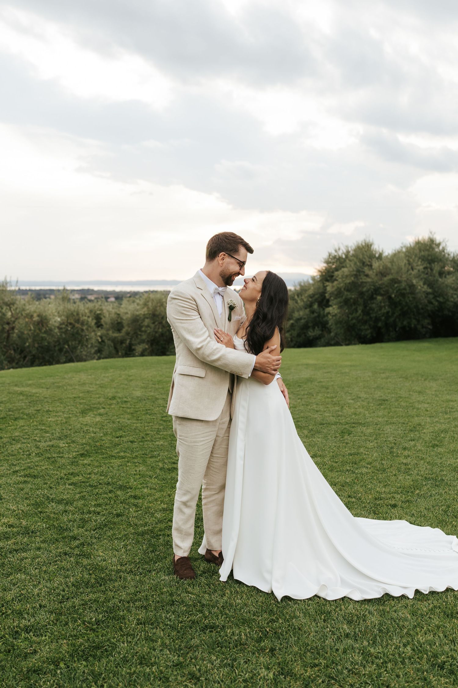 A bride and groom share a romantic embrace on a lush green lawn with trees and cloudy sky backdrop.