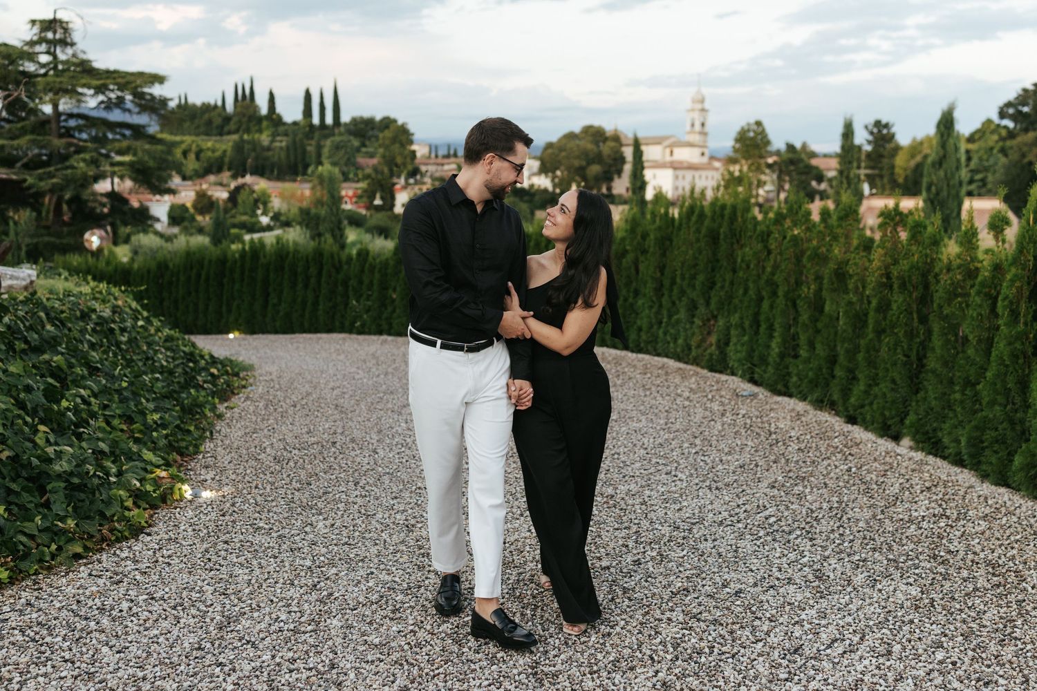 A couple embraces on a gravel path surrounded by manicured hedges and cypress trees in an elegant garden setting.