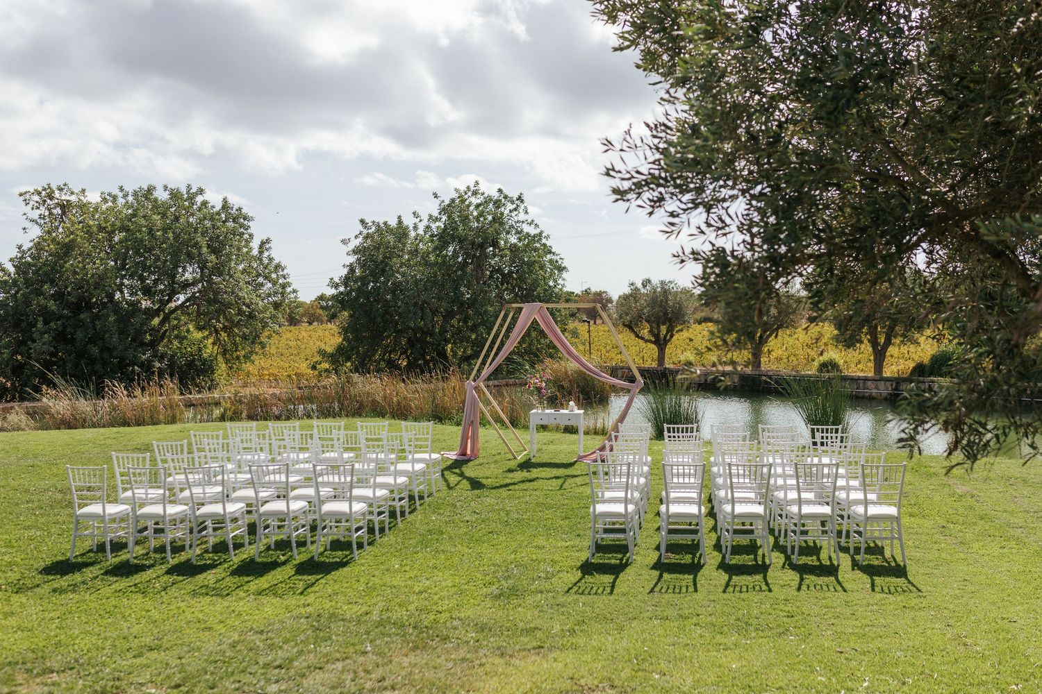 Outdoor wedding ceremony setup with white chairs and wooden arch overlooking a serene pond.