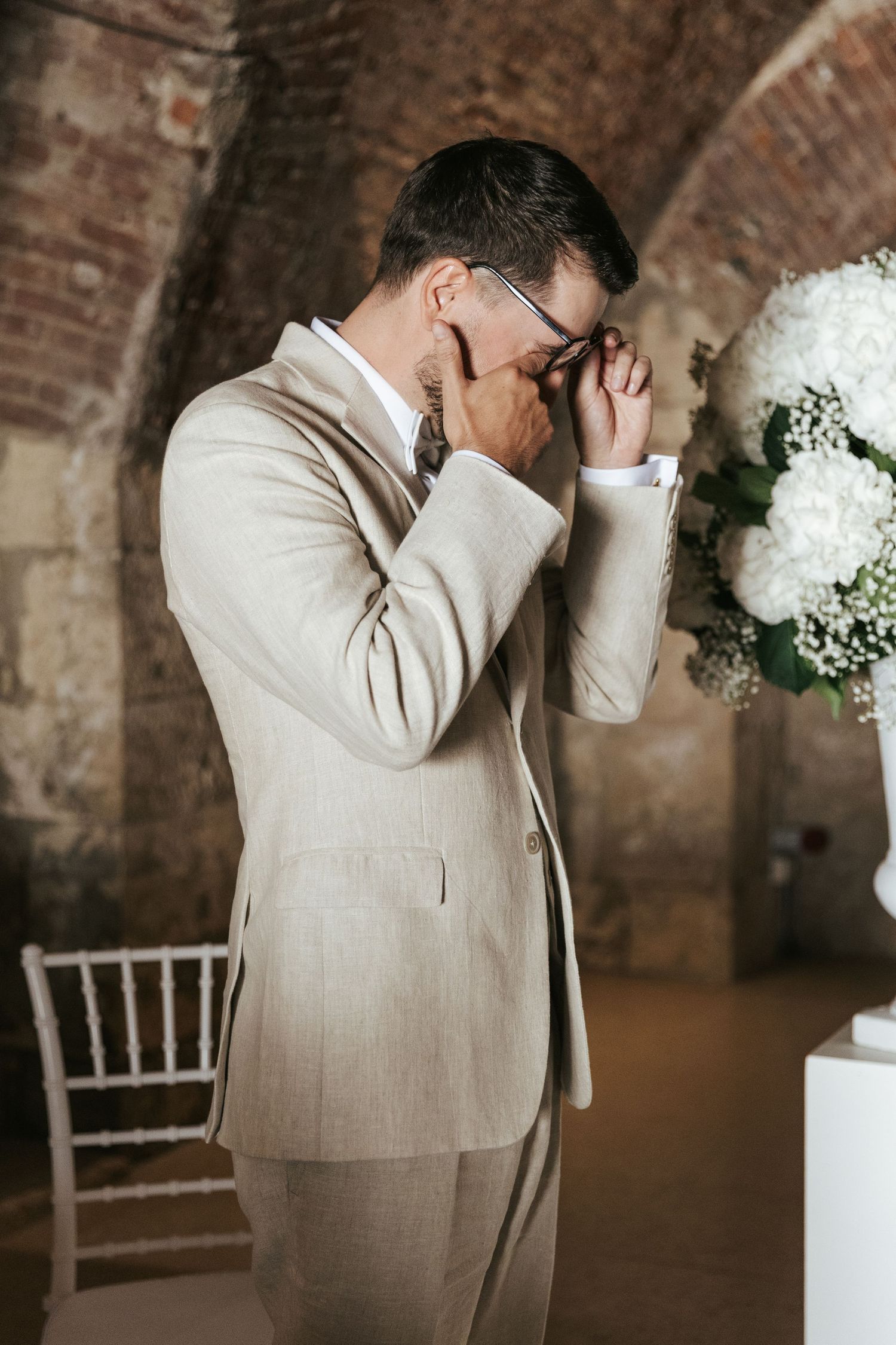 Emotional groom in beige suit wipes tears during wedding ceremony in brick venue.