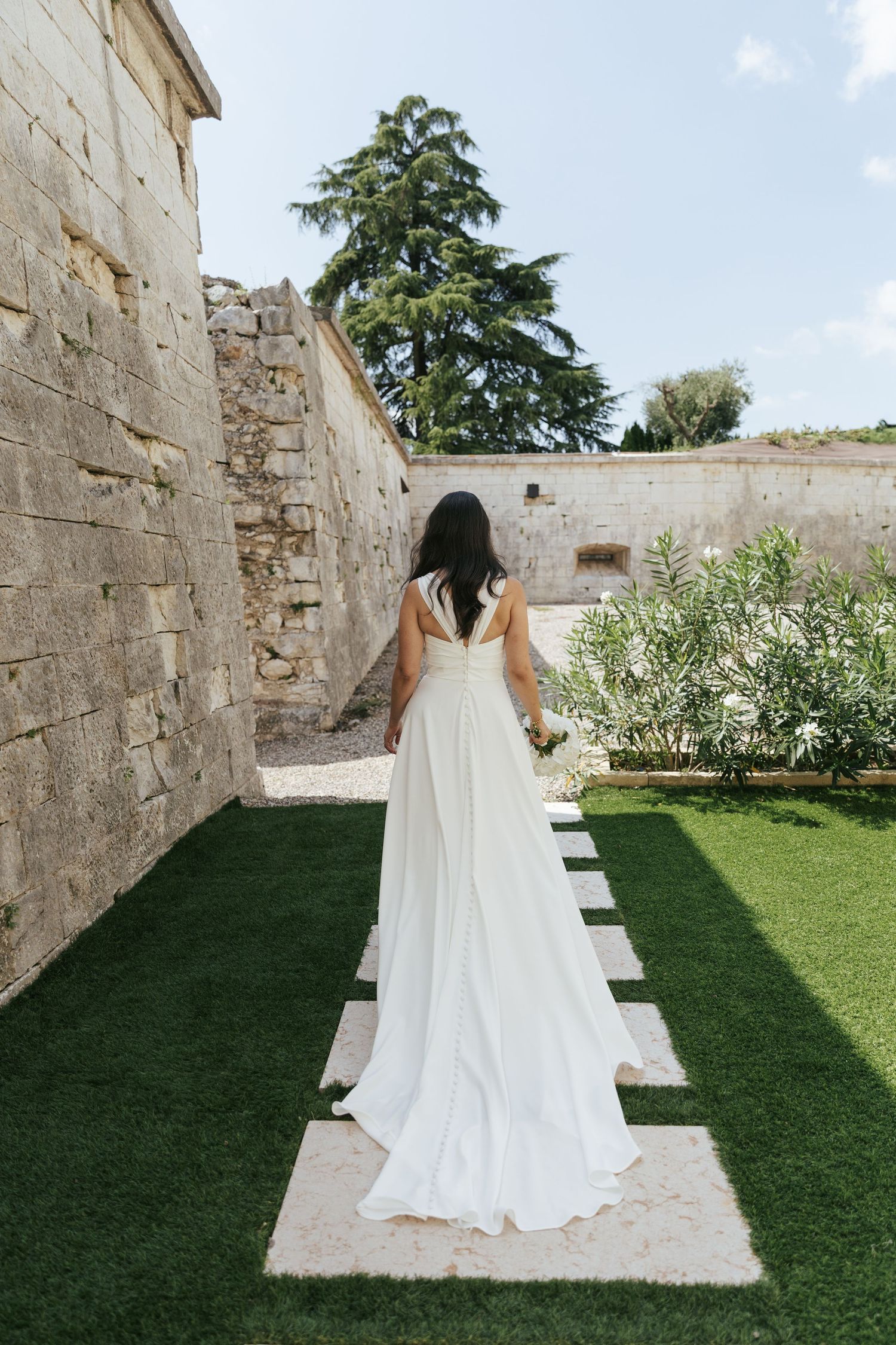 A wedding dress with flowing train photographed from behind on a garden path with stone walls.
