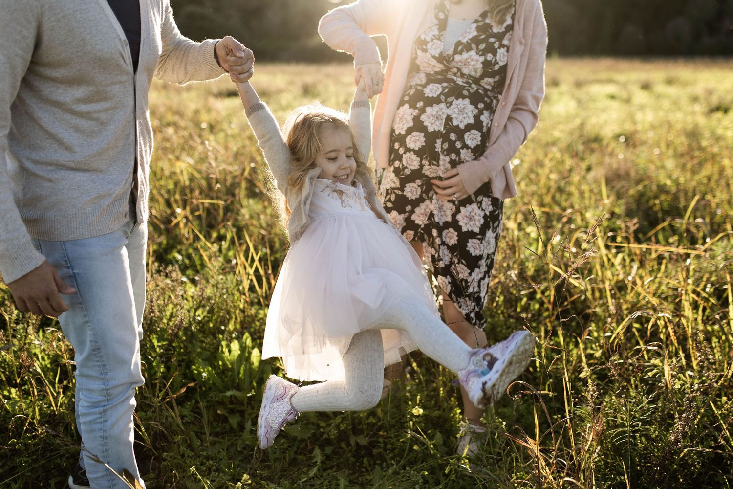 Family playing together in a sunlit meadow, little one jumping in a white dress while holding hands with adults.