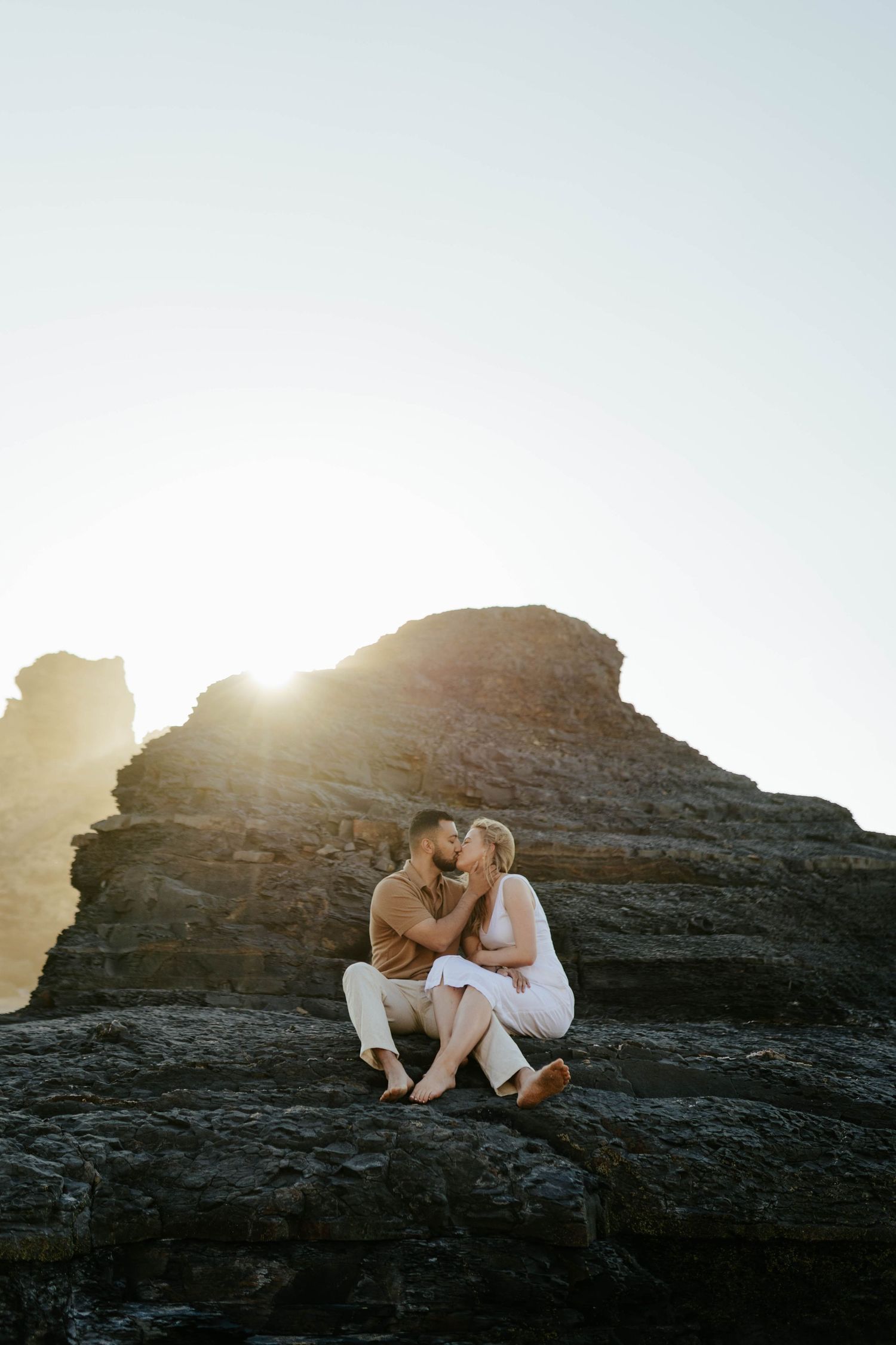 A romantic moment captured at sunset with two people embracing on coastal rocks as sunlight streams behind them.