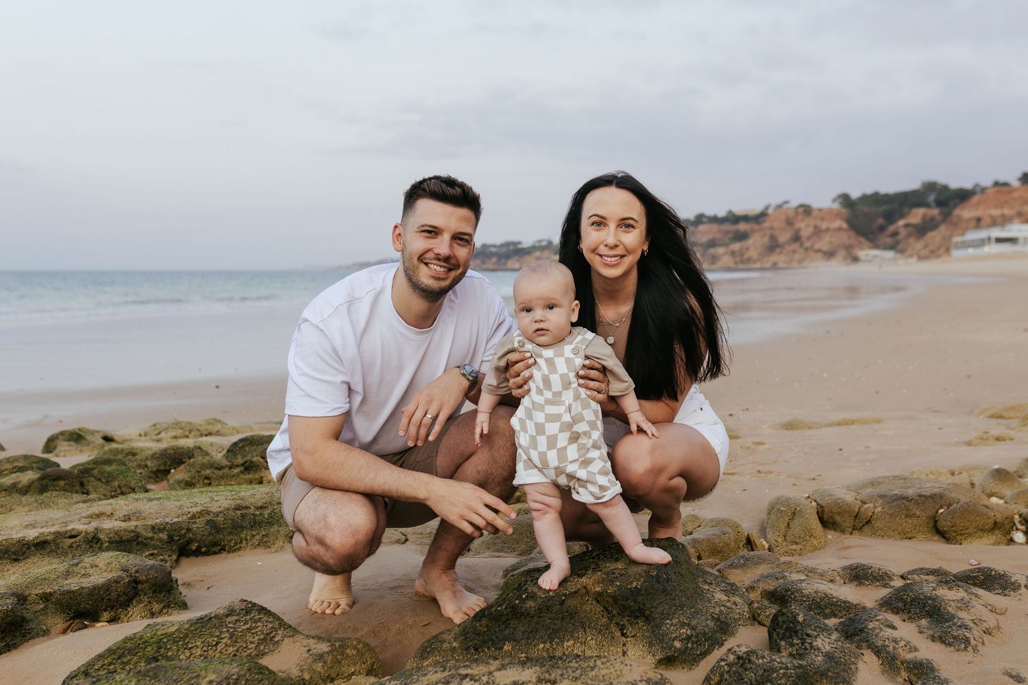 A young family poses together on rocky beach terrain with ocean waves and cliffs in the background.