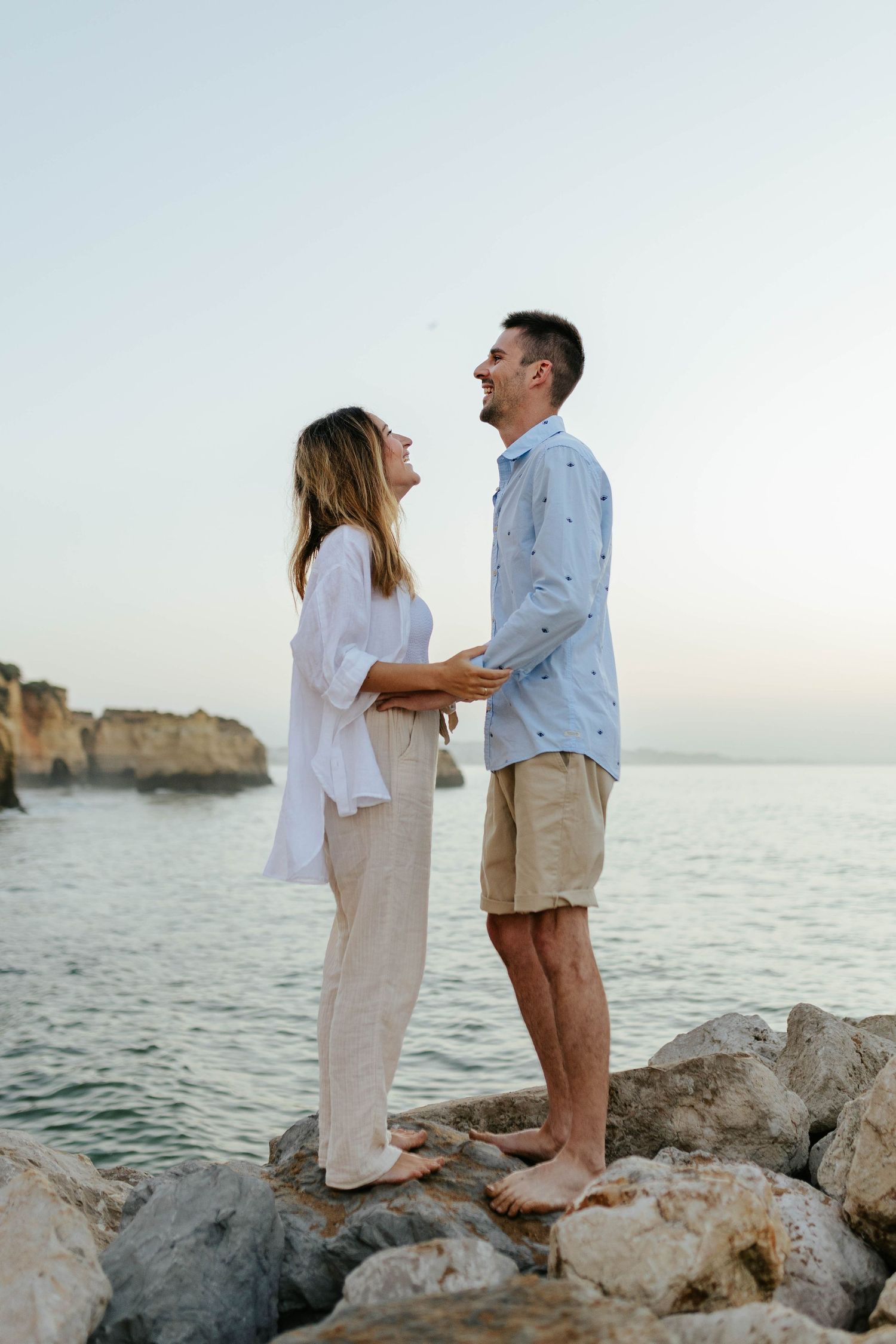 A couple shares a tender moment while standing on rocky coastal formations at sunrise. Praia Dona Ana, Lagos, Algarve, Portugal.