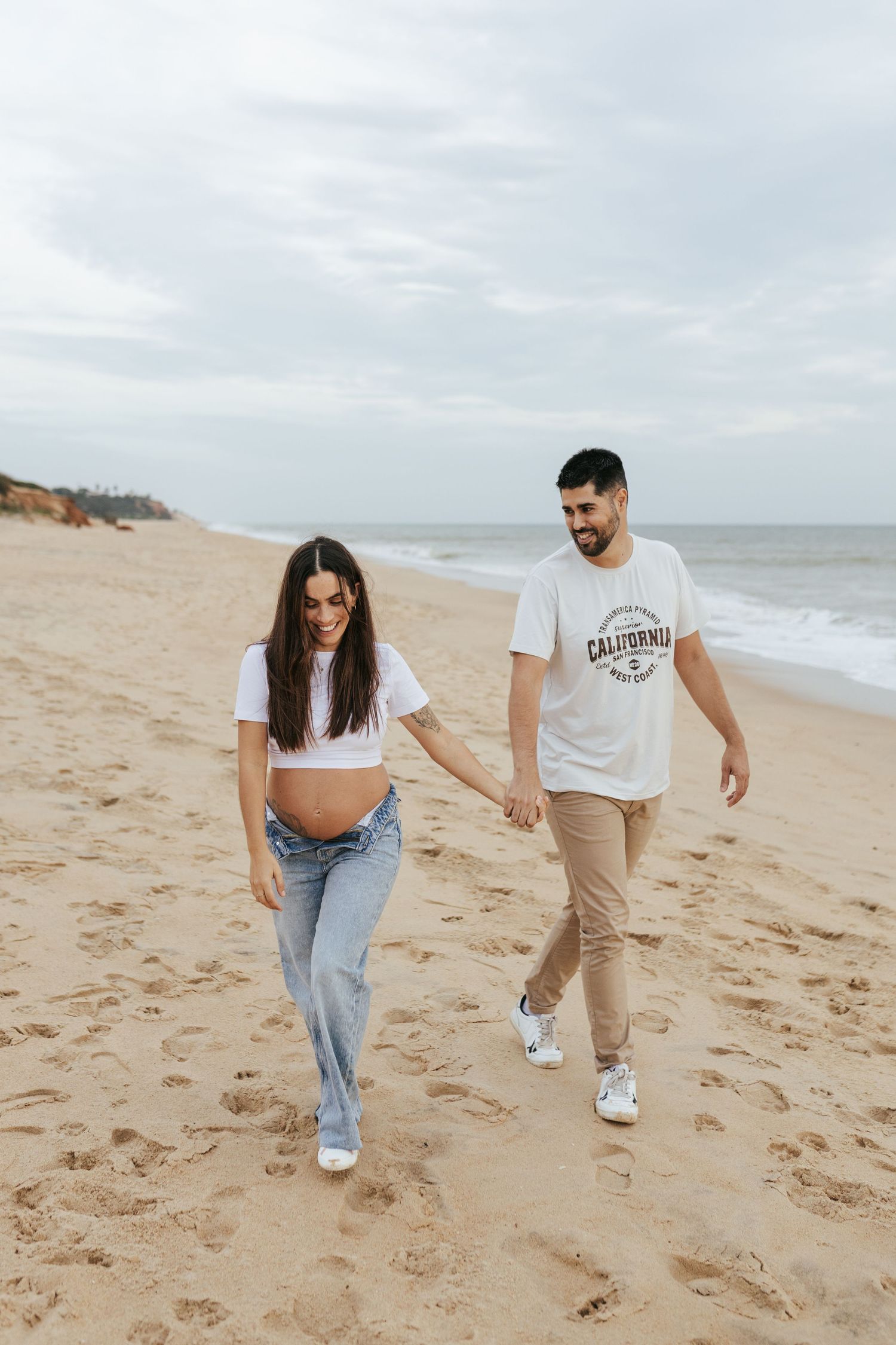 A couple walking together and smiling on a sandy beach with ocean waves and cloudy sky in the background.