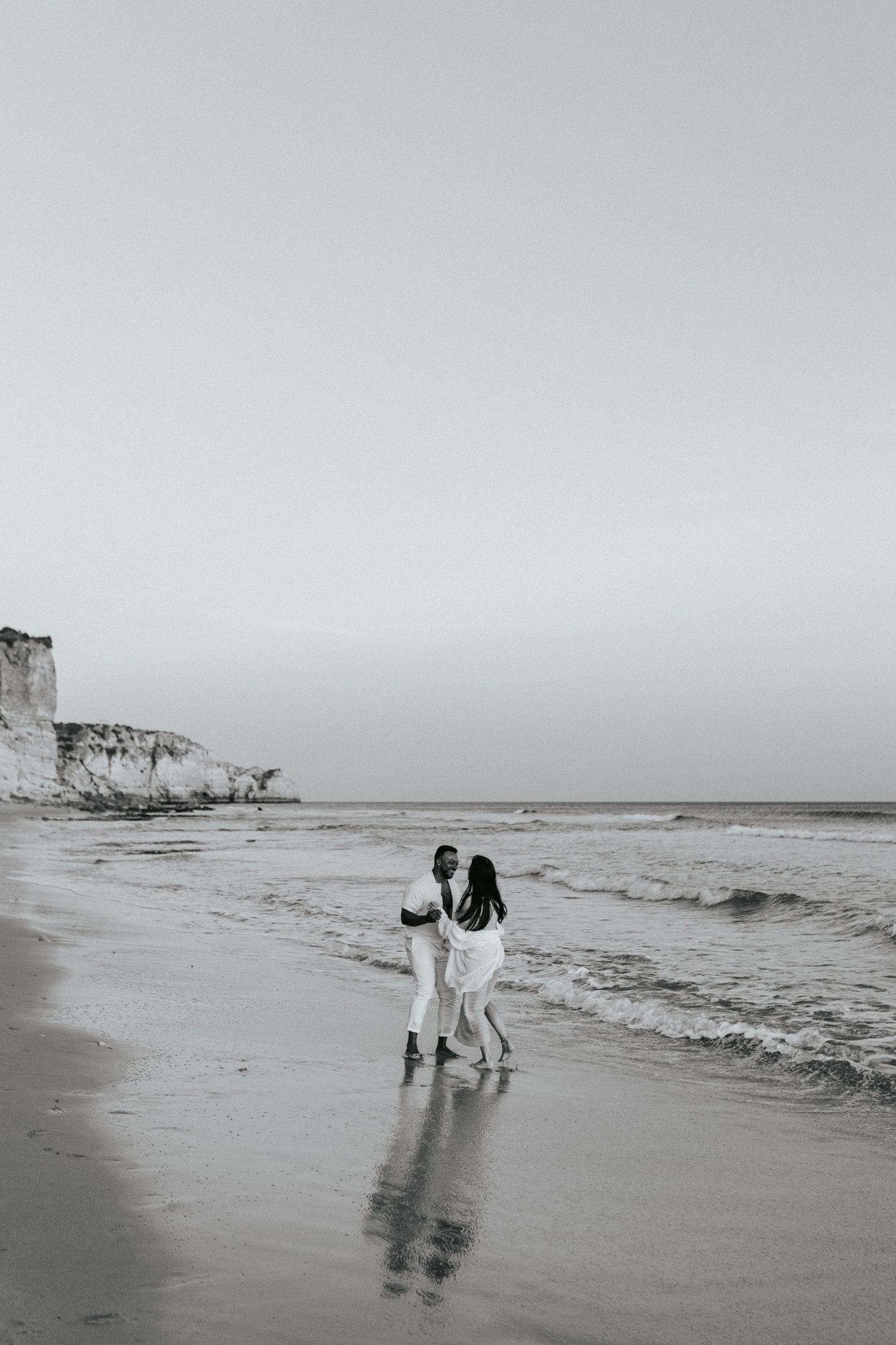 Two figures walking together along a misty beach shoreline with dramatic coastal cliffs in the background.