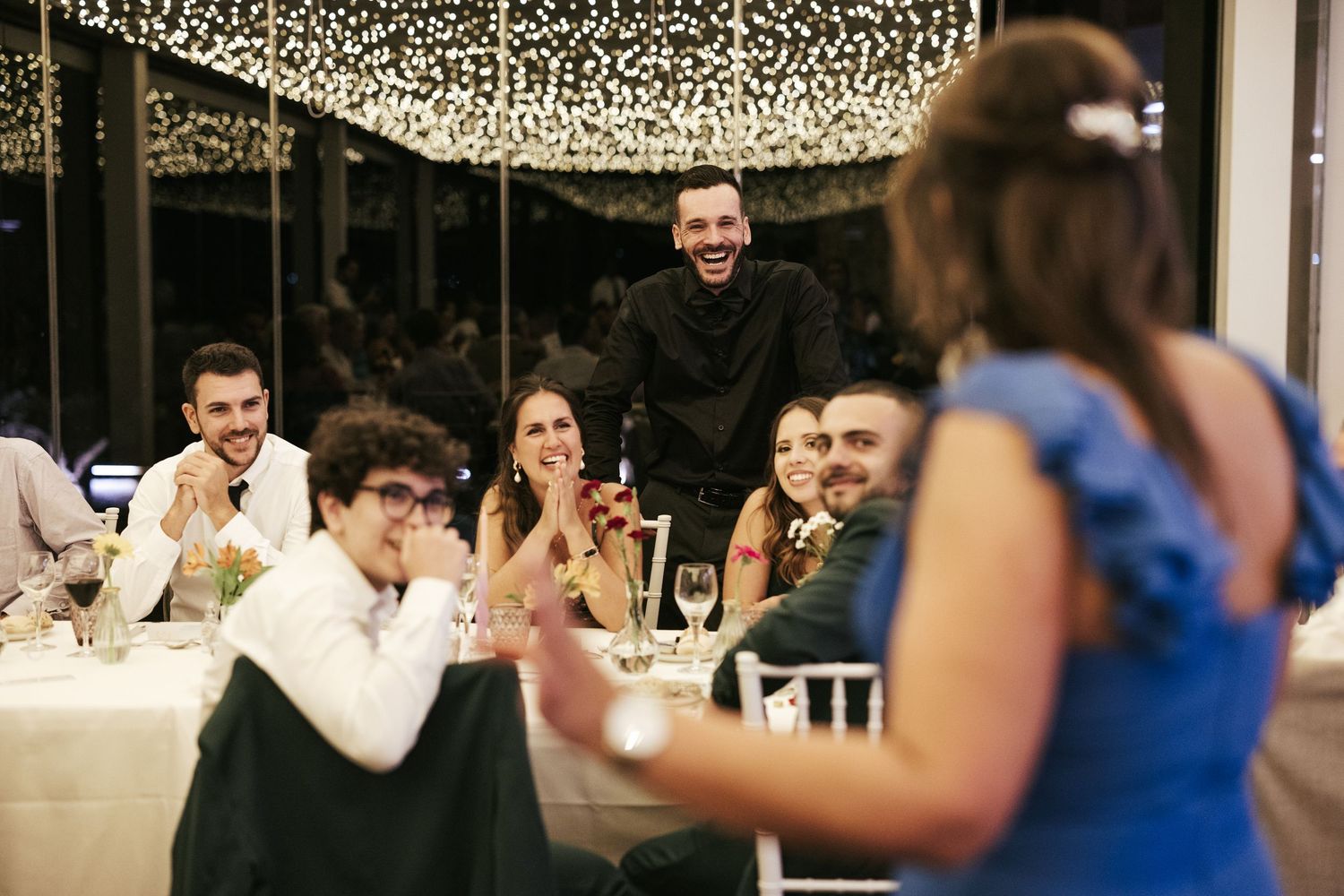 Wedding guests react with laughter during reception under twinkling ceiling lights.