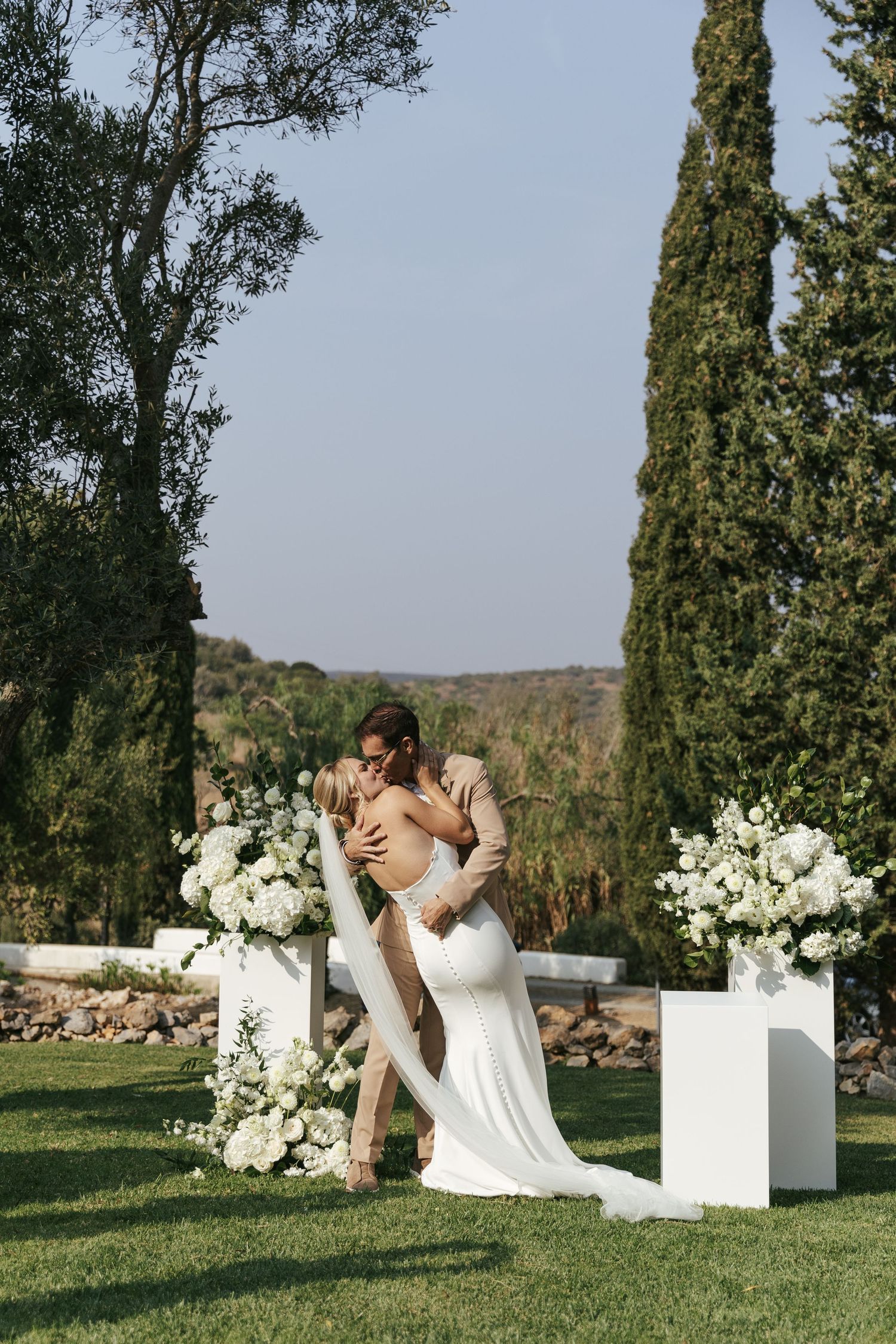 Romantic outdoor wedding ceremony with white floral pillars and cypress trees in a scenic landscape.