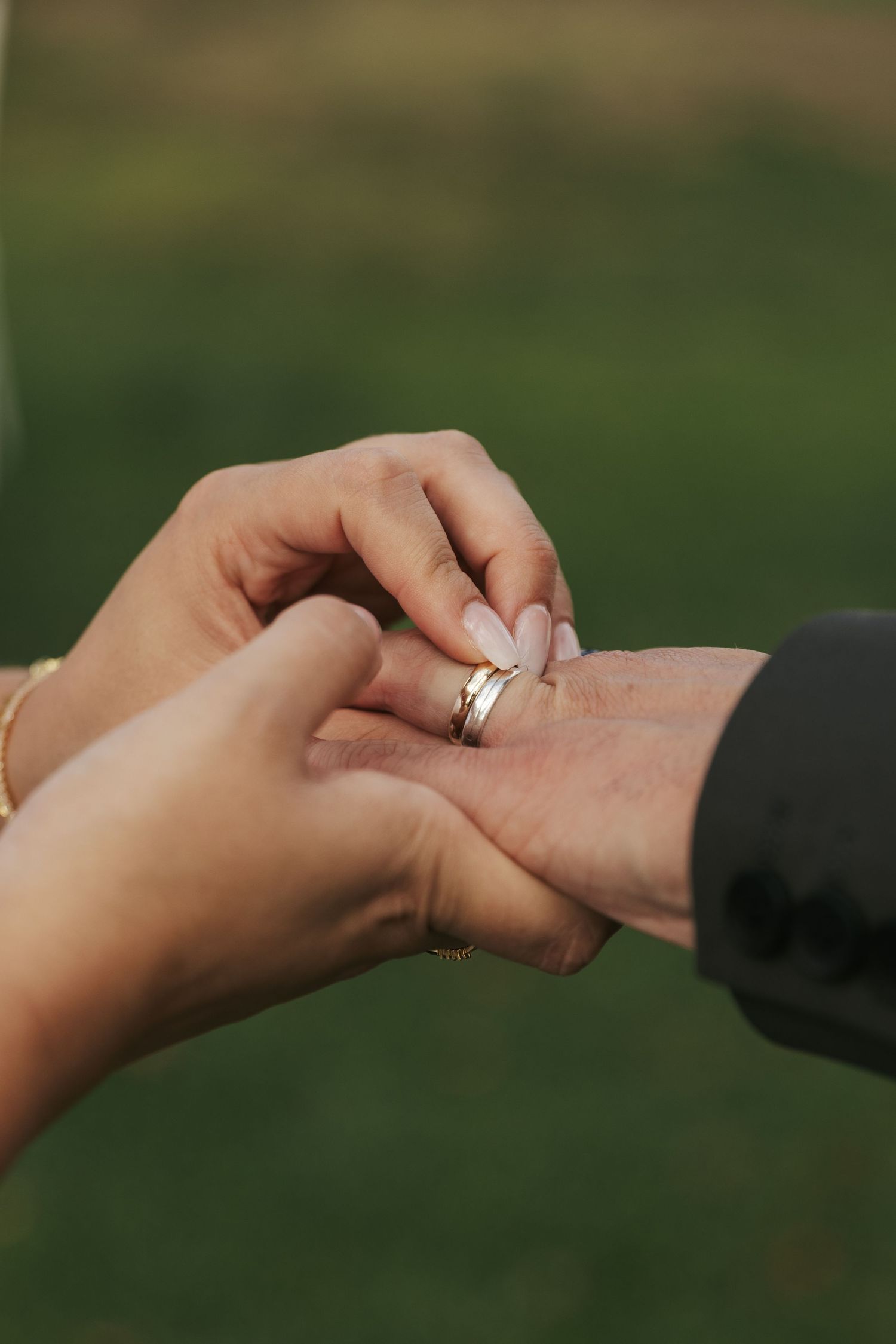 Close-up of hands exchanging wedding rings during an outdoor ceremony against a green background.