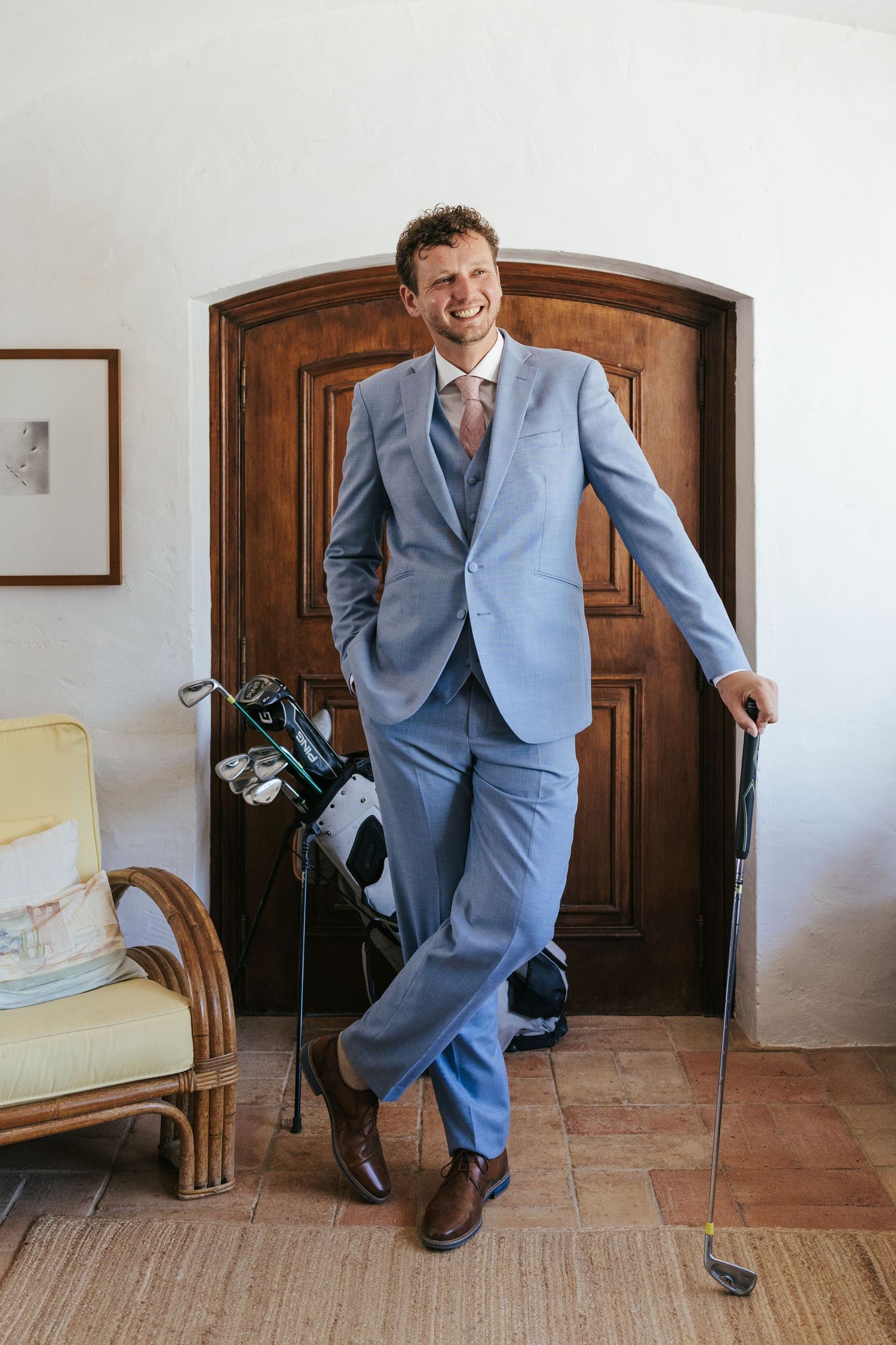 Man in light blue suit holding golf club, smiling by wooden door with golf bag, wearing brown dress shoes.