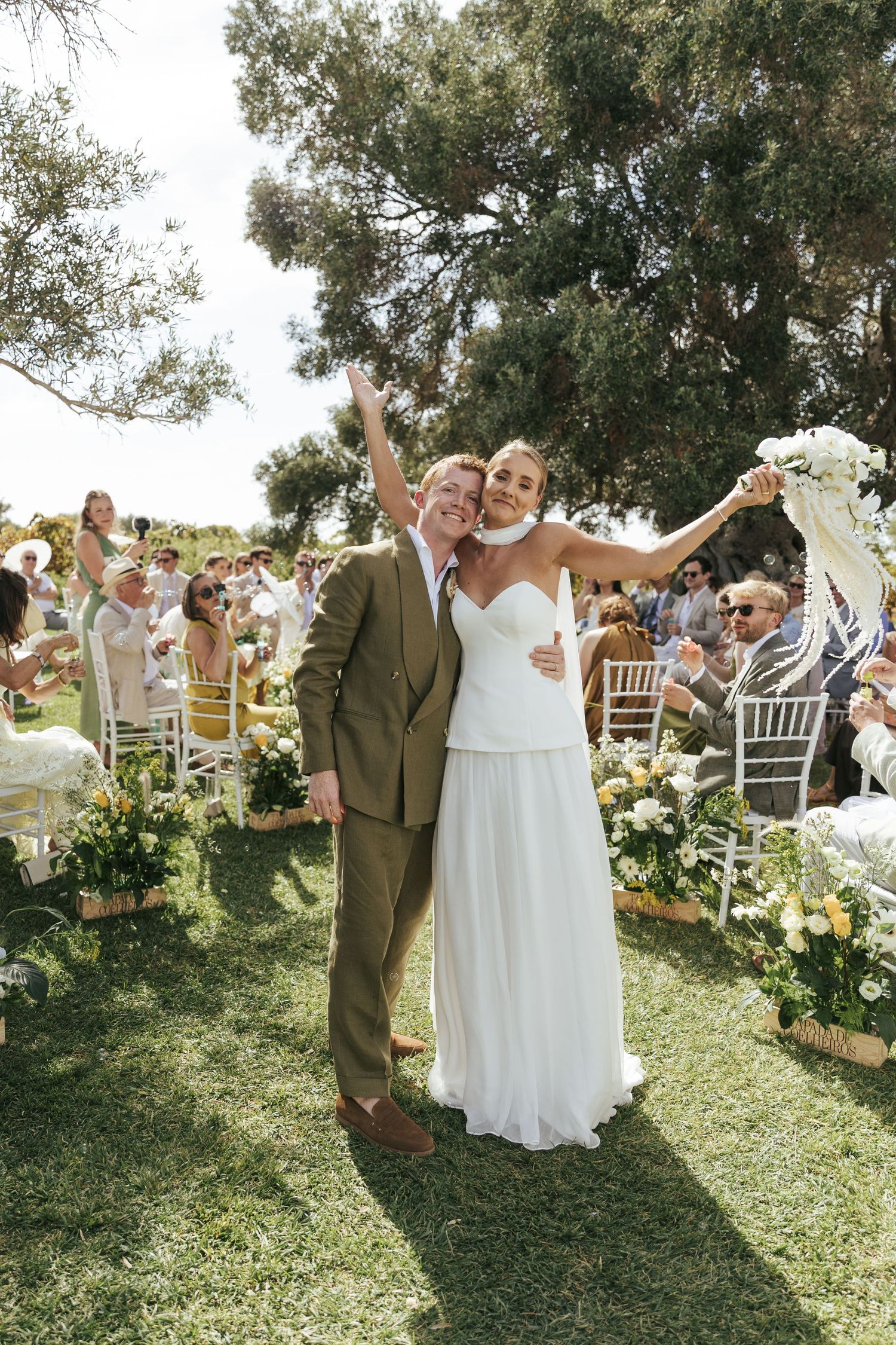 Joyful newlyweds celebrate after outdoor wedding ceremony, bride in white gown and groom in olive suit, guests seated on lawn.