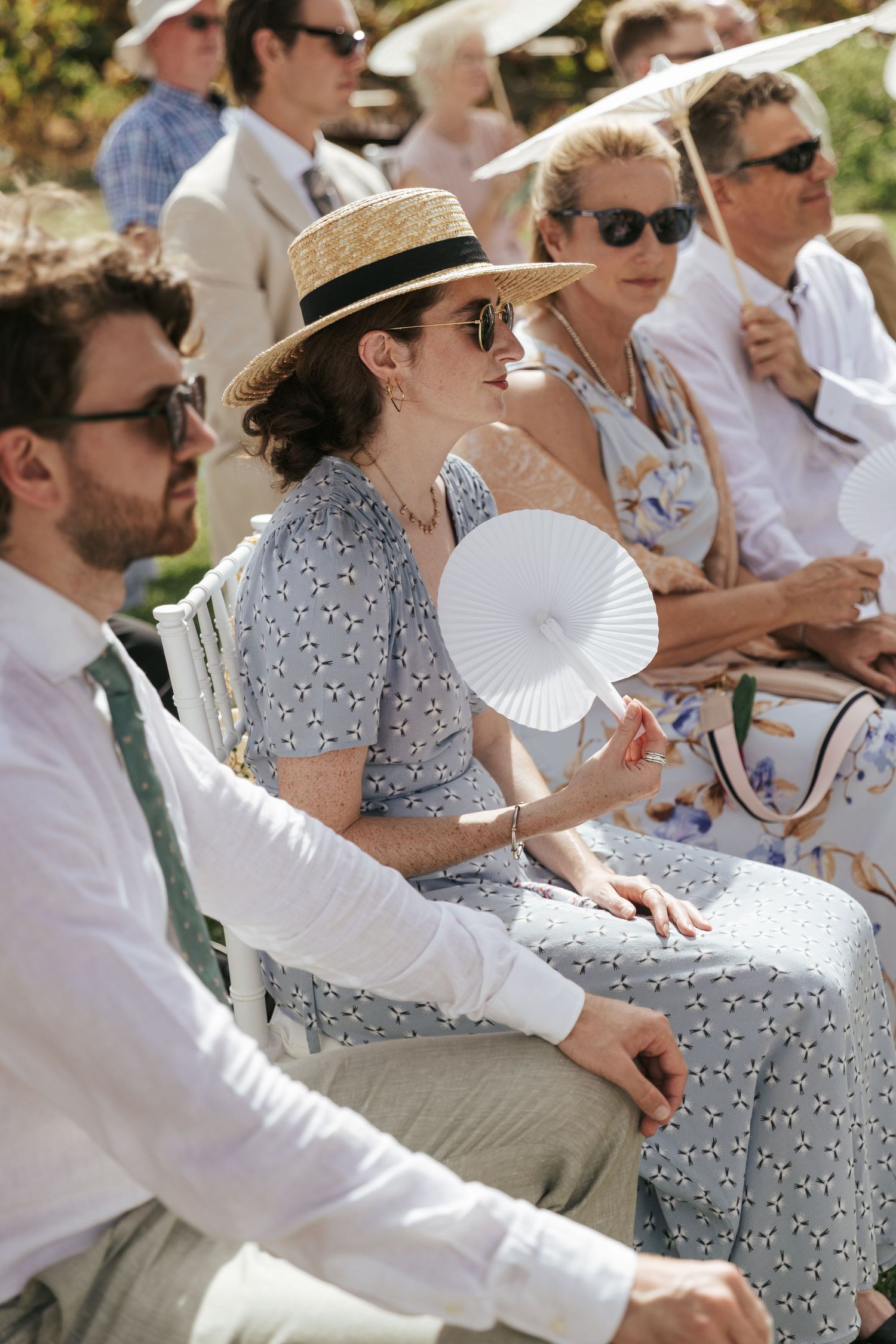 Woman in straw hat and sunglasses holding white paper fan at outdoor summer wedding ceremony.