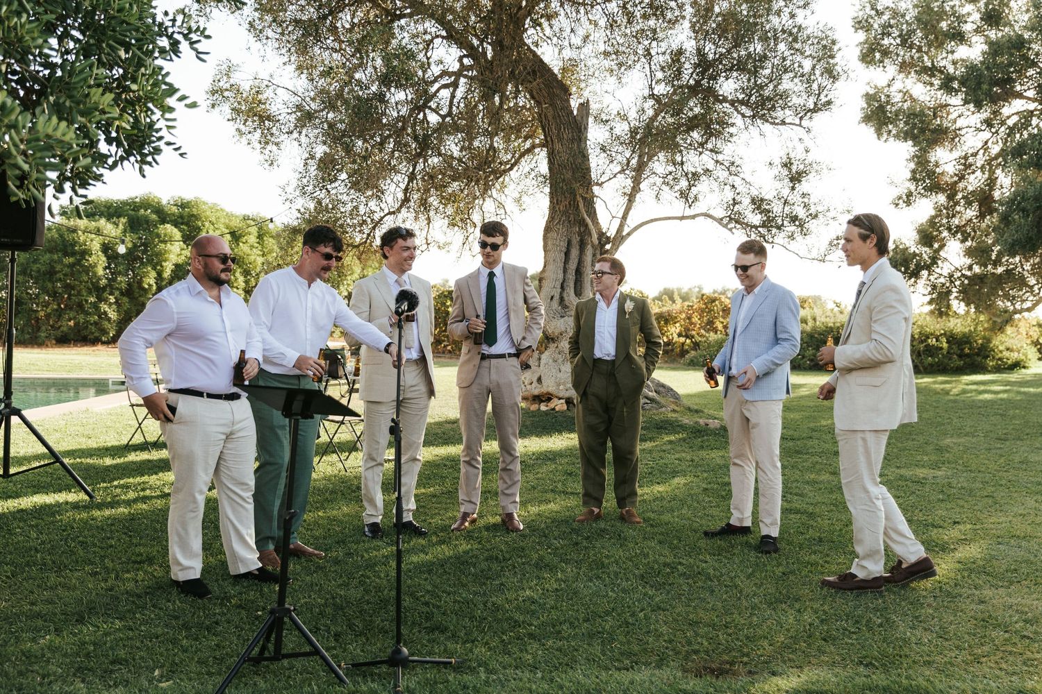 Group of men performing or speaking at an outdoor wedding ceremony under an olive tree.