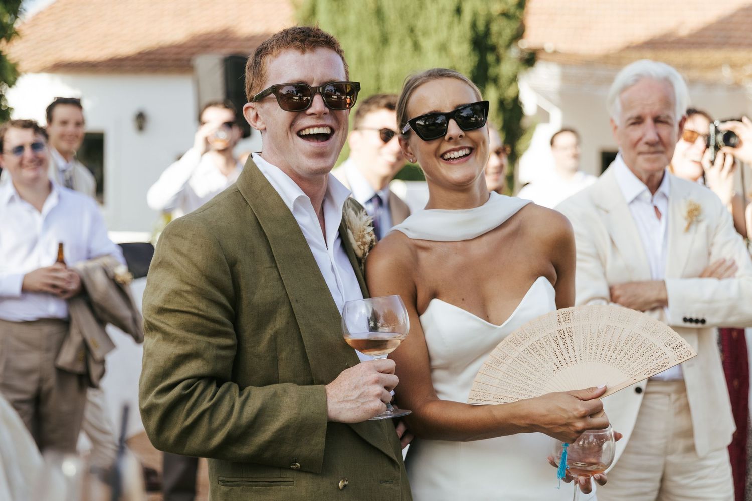 Laughing bride in white dress and groom in olive suit wearing sunglasses at outdoor wedding reception.