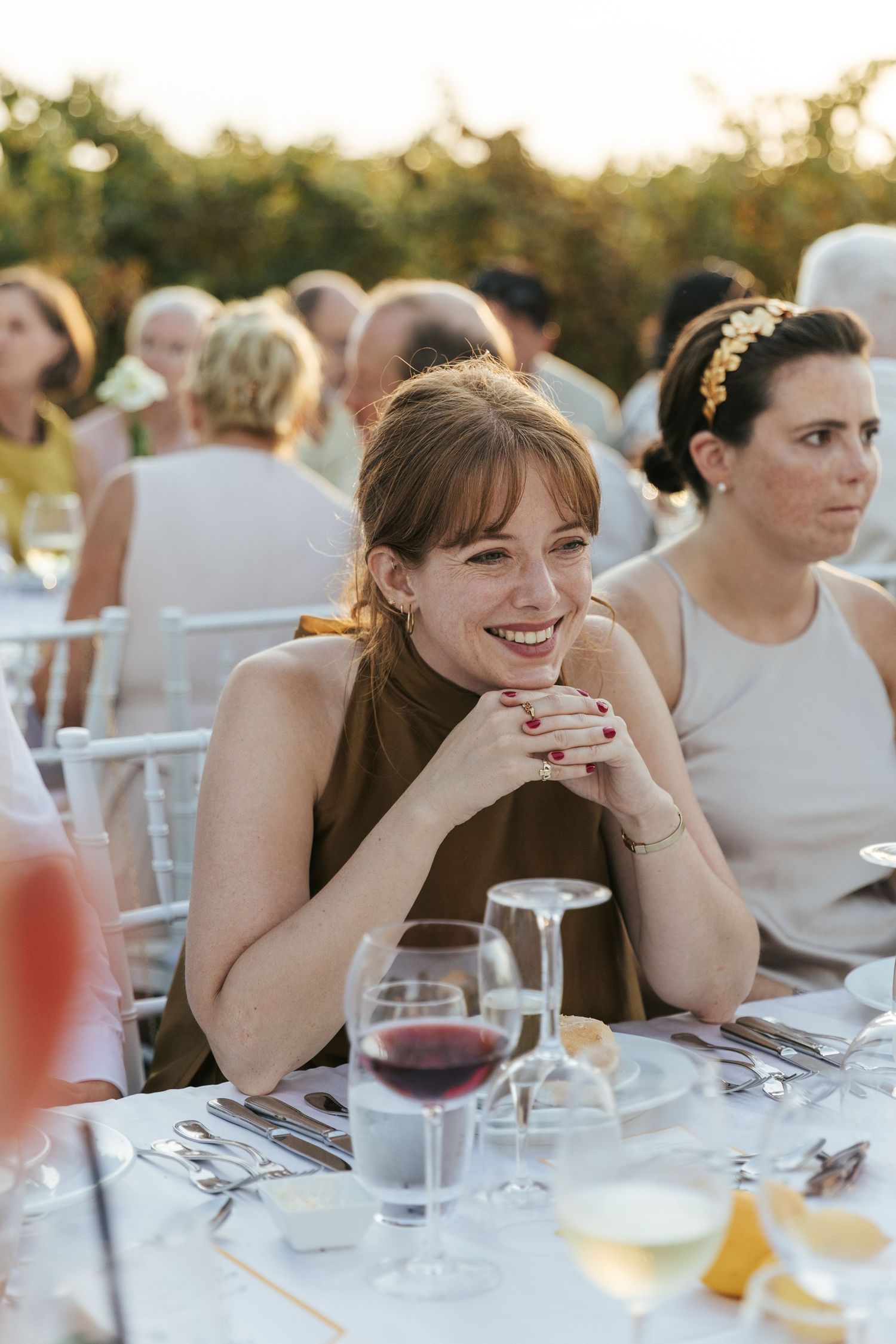 Smiling woman in brown dress enjoying outdoor wedding reception dinner with wine glasses on table.