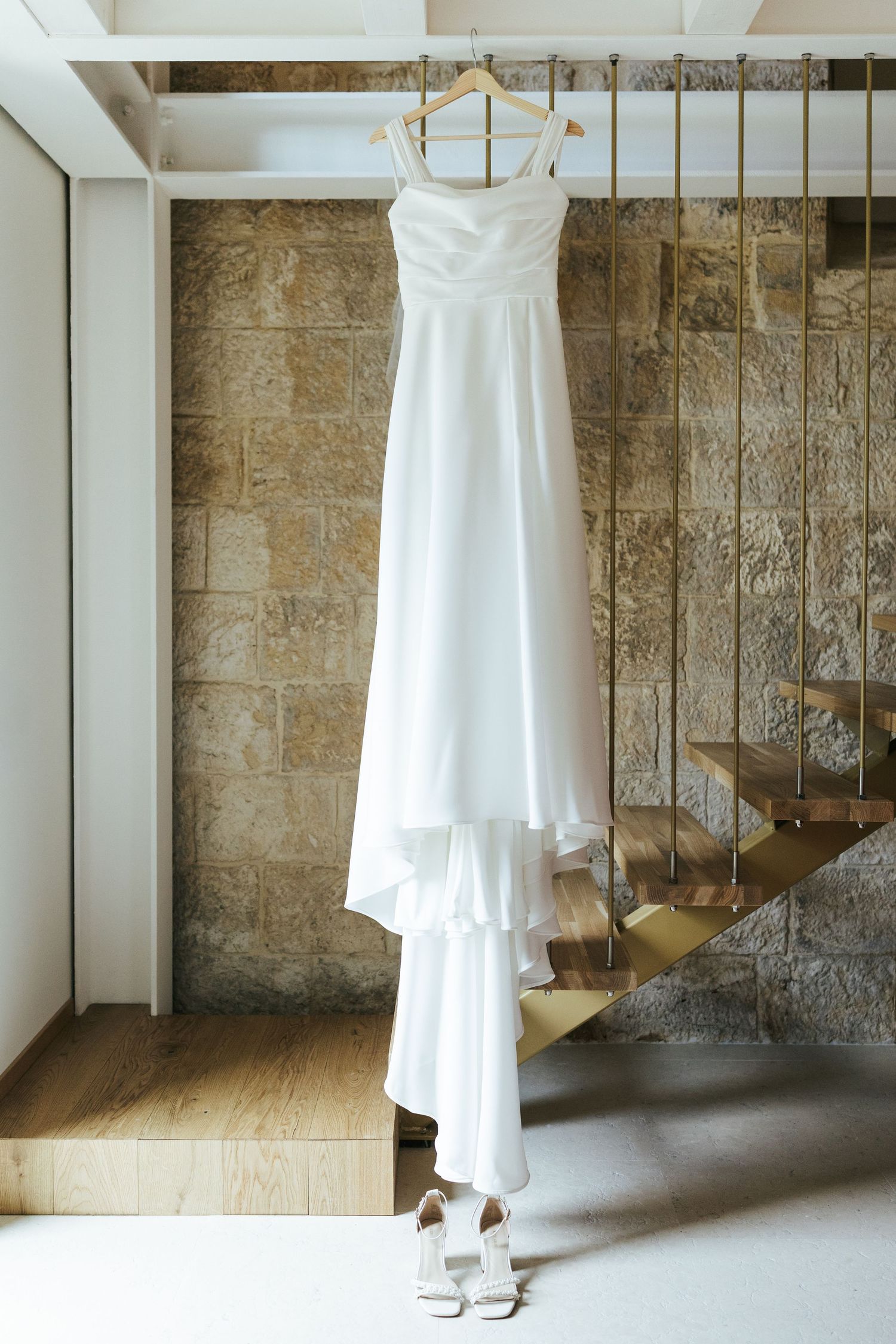 A white wedding dress hangs elegantly against a stone wall with white pillars and wooden stairs.