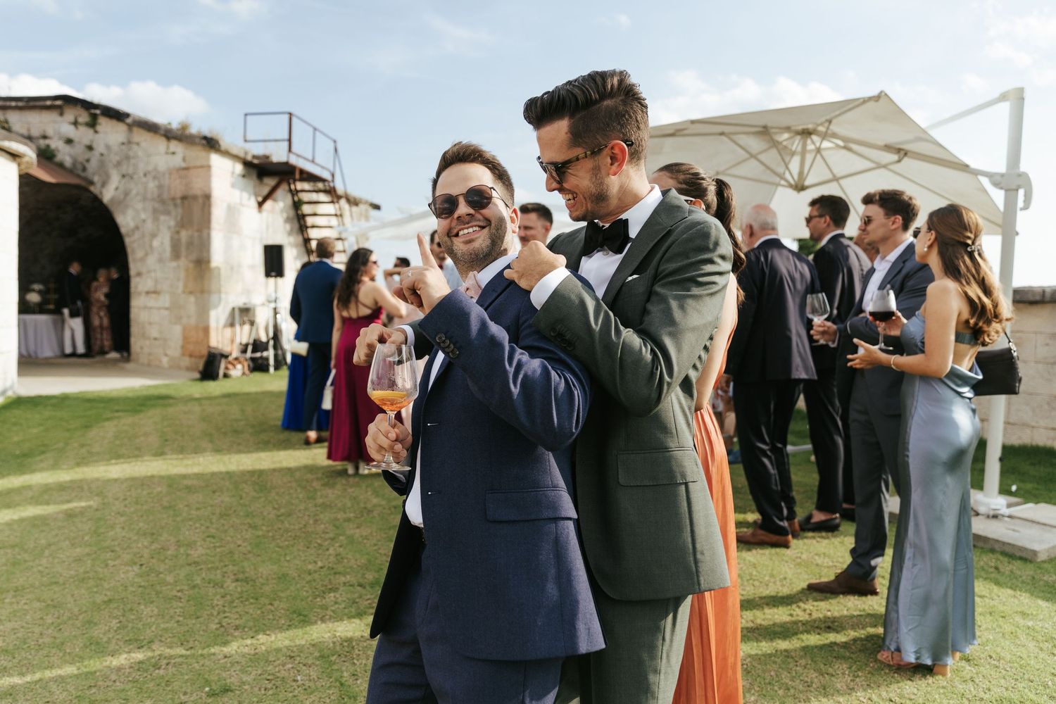 Wedding guests socialize during an outdoor reception near a stone building.