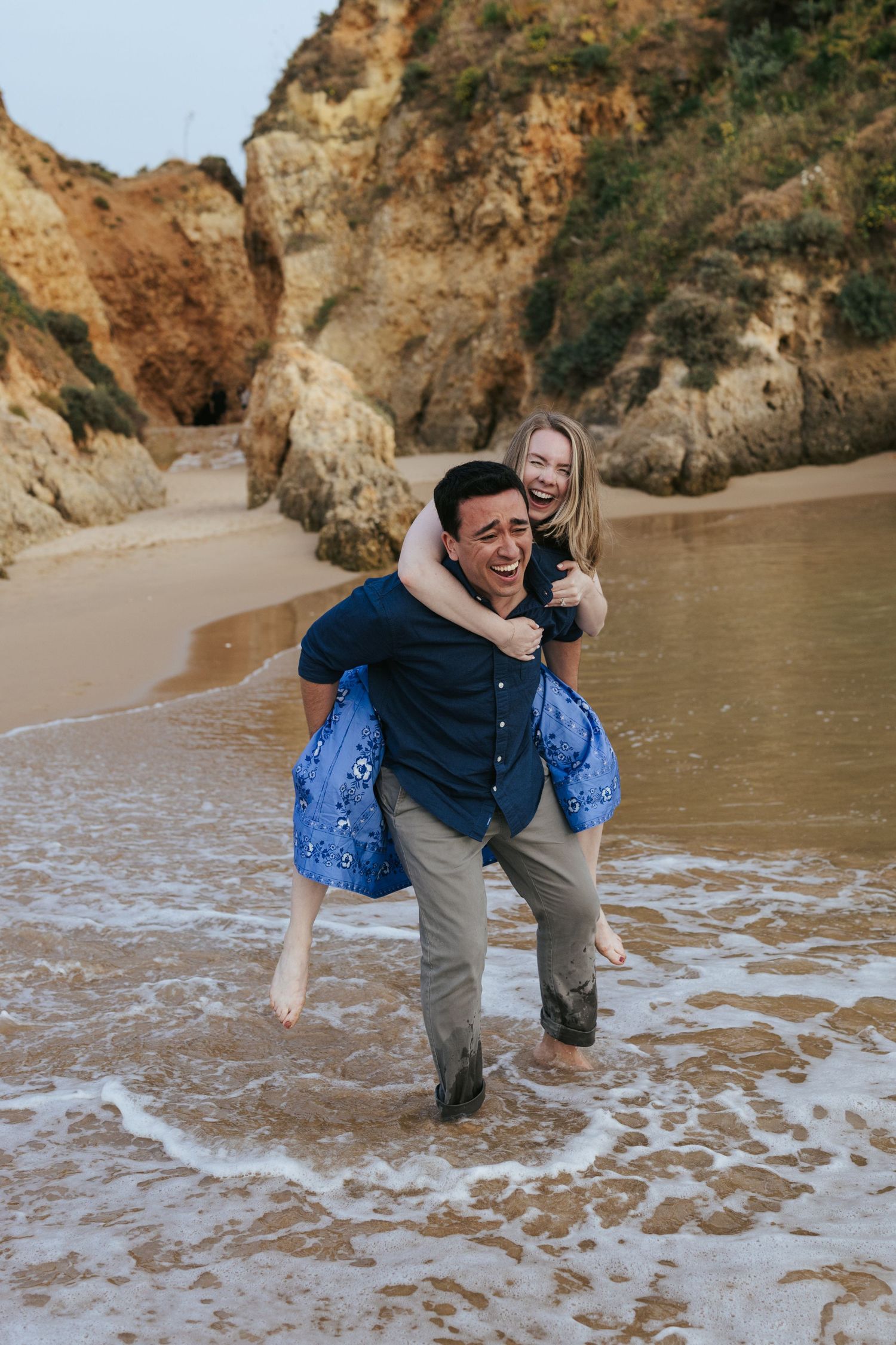 A man gives a laughing woman a piggyback ride on a sandy beach with rocky cliffs in the background.