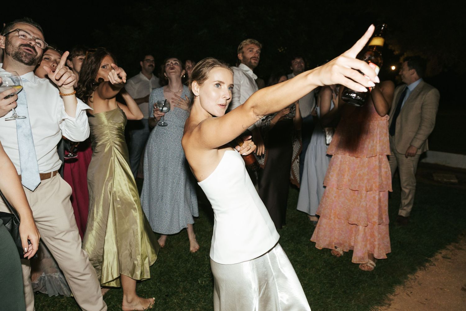 A woman in a white satin dress dances energetically at an outdoor wedding reception at night.