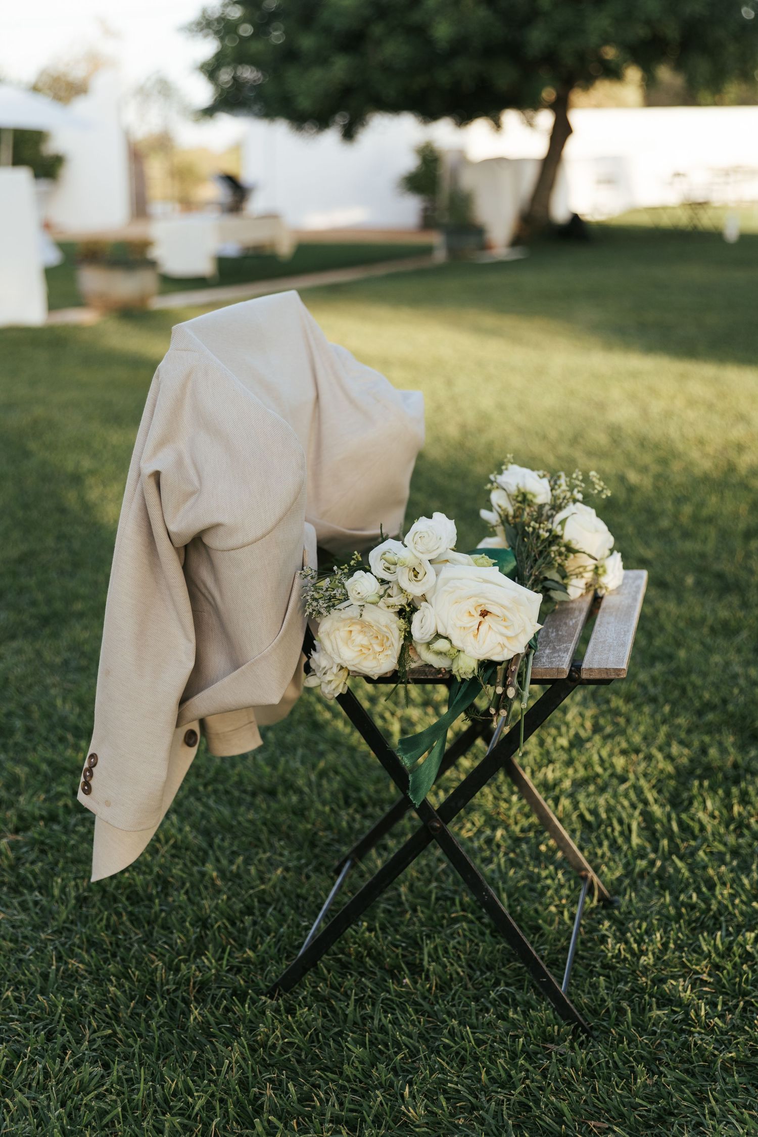 White rose bridal bouquet resting on folding chair with beige blazer on green lawn.