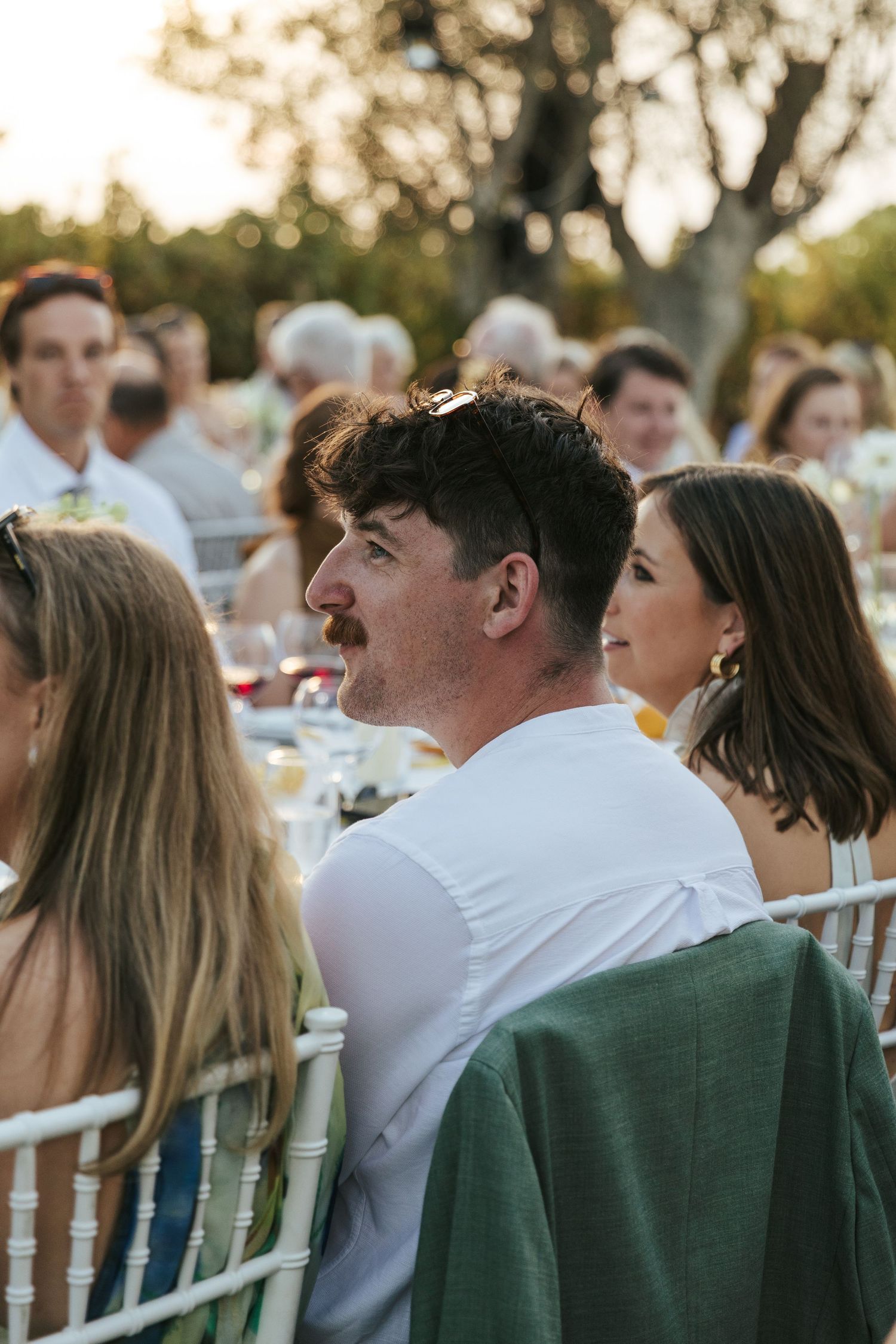 Man with mustache in white shirt and green jacket seated at outdoor wedding reception at sunset.