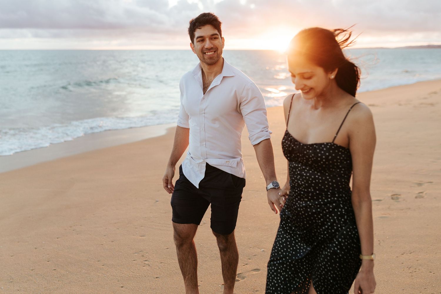 A couple walks along a sandy beach at sunset with ocean waves and glowing sunlight in the background.