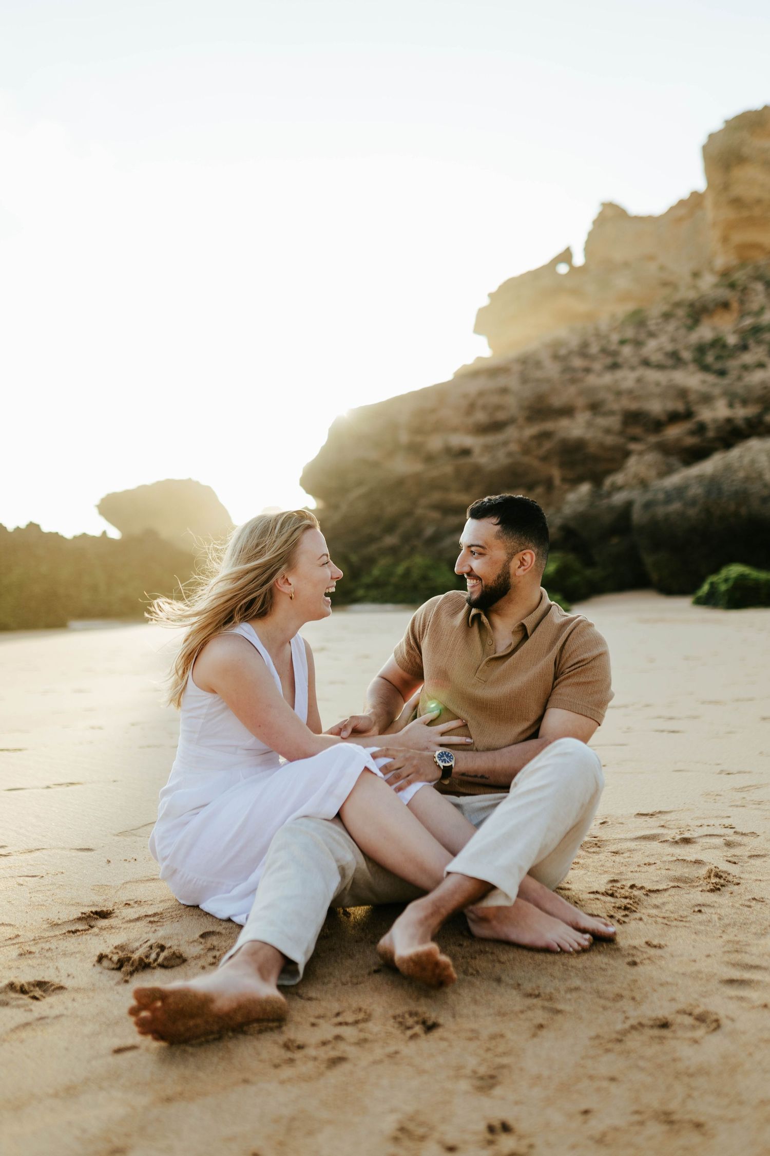 A couple sitting on a sandy beach at sunset laughing together with cliffs in the background.