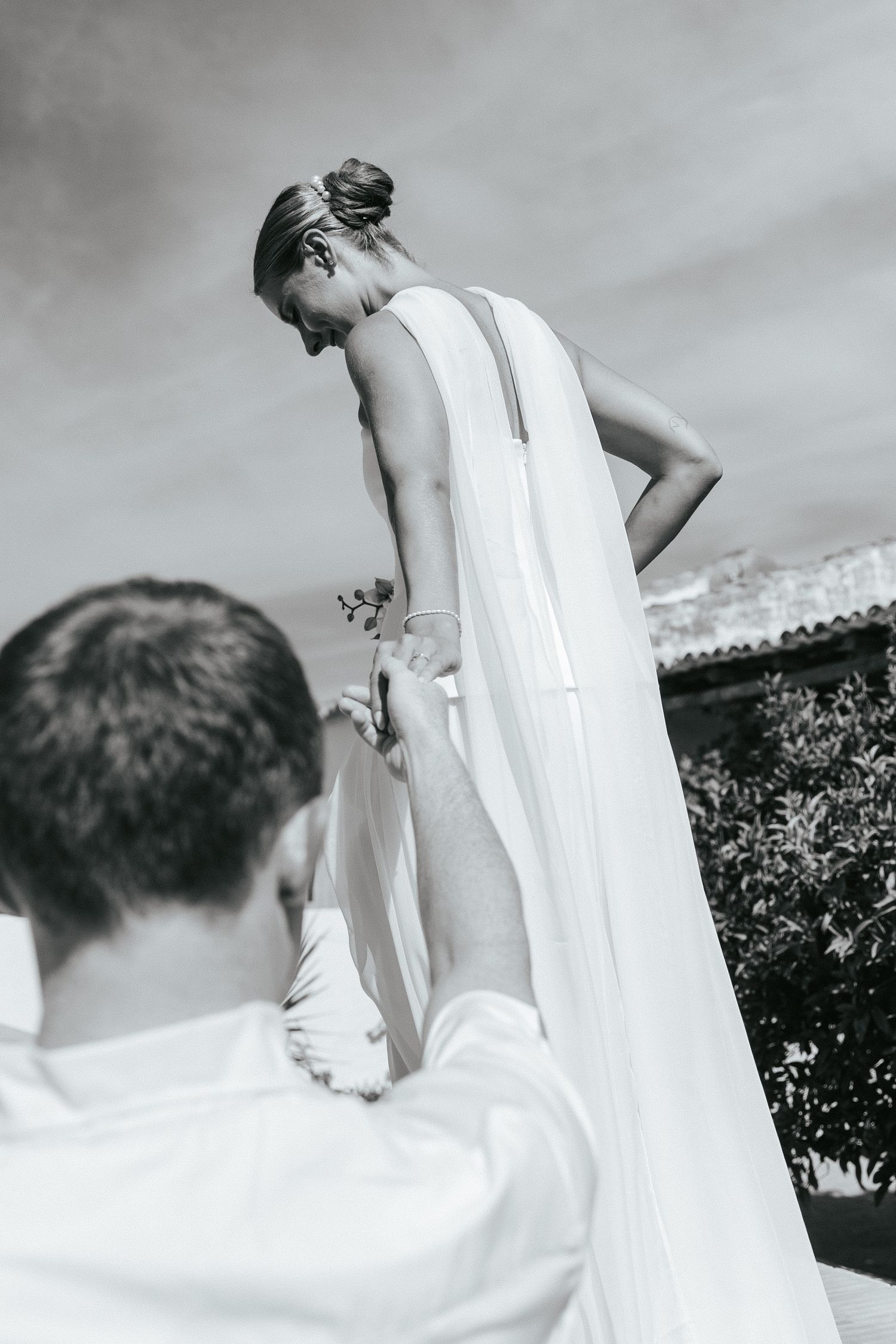 Black and white wedding photo showing a person adjusting a long white dress train outdoors under cloudy skies.