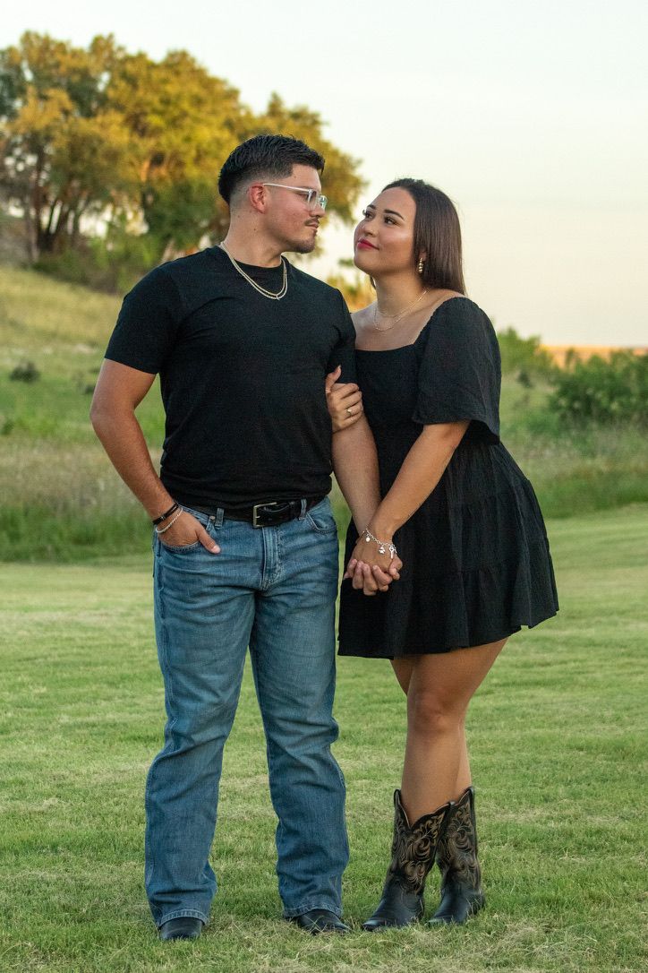 A romantic couple wearing matching black tops stands together on a grassy field during golden hour.