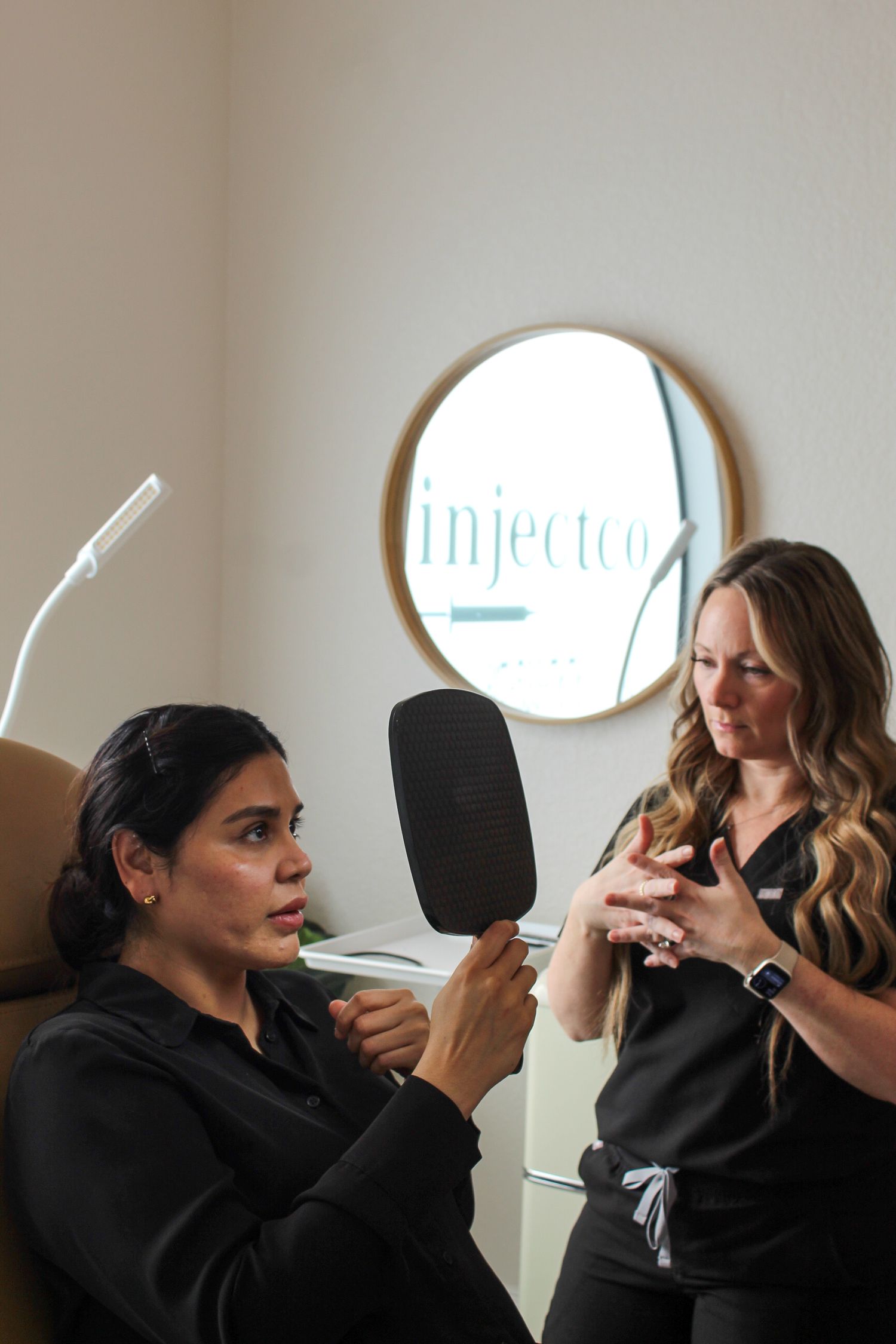 Two beauty professionals in black attire having a consultation at an aesthetics clinic with Injector sign in background.