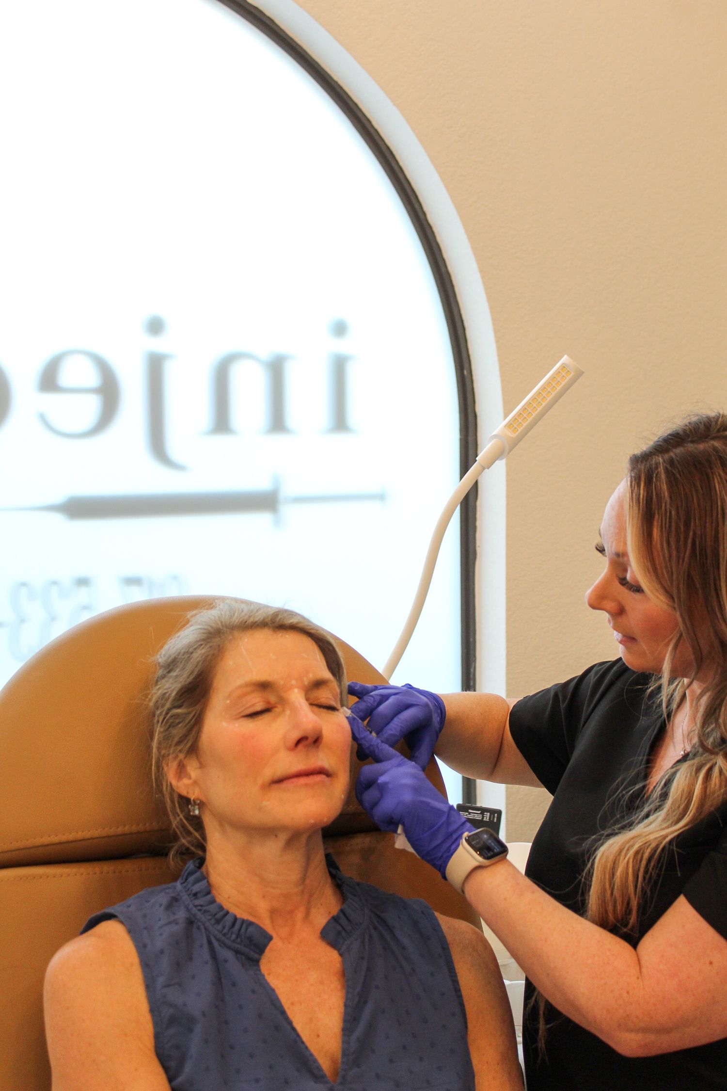 An aesthetician wearing purple gloves performs a cosmetic procedure on a client in a medical spa setting.