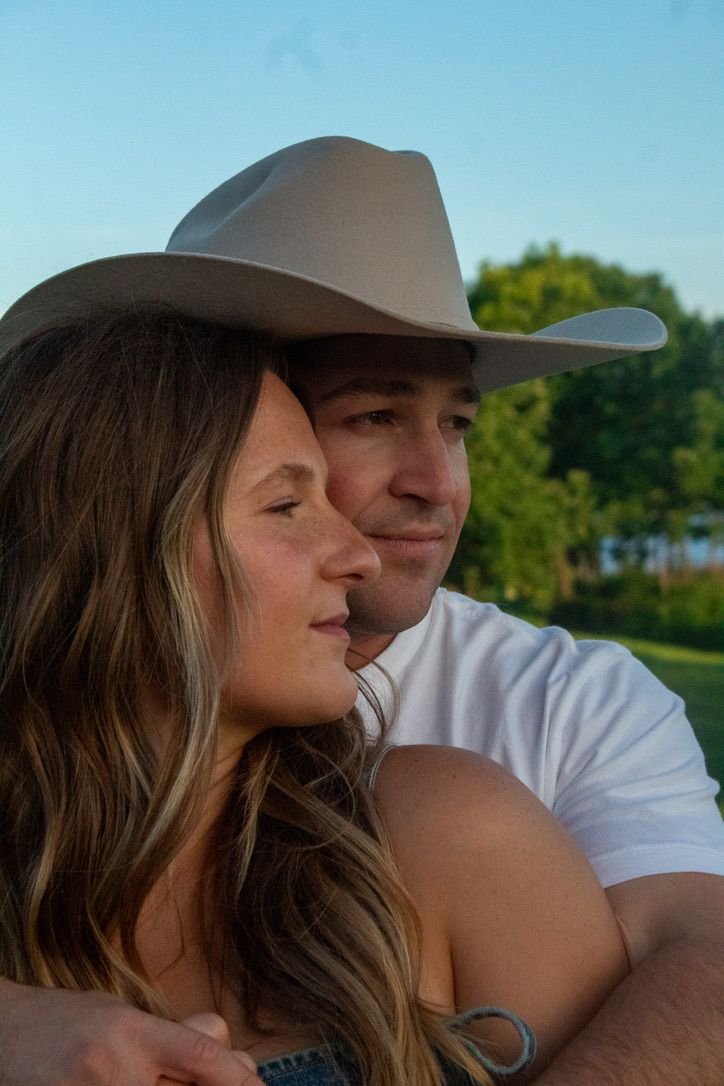 A couple embraces outdoors during golden hour, with one wearing a gray cowboy hat and white t-shirt.