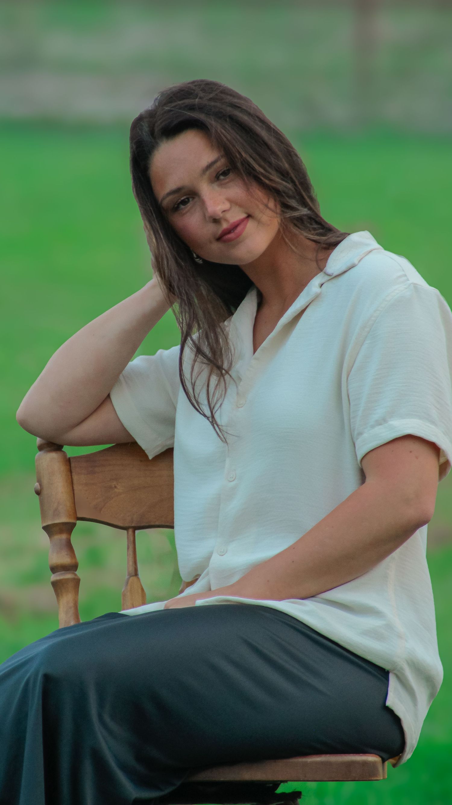 A relaxed outdoor portrait of someone in white shirt sitting on wooden chair against green grass background.