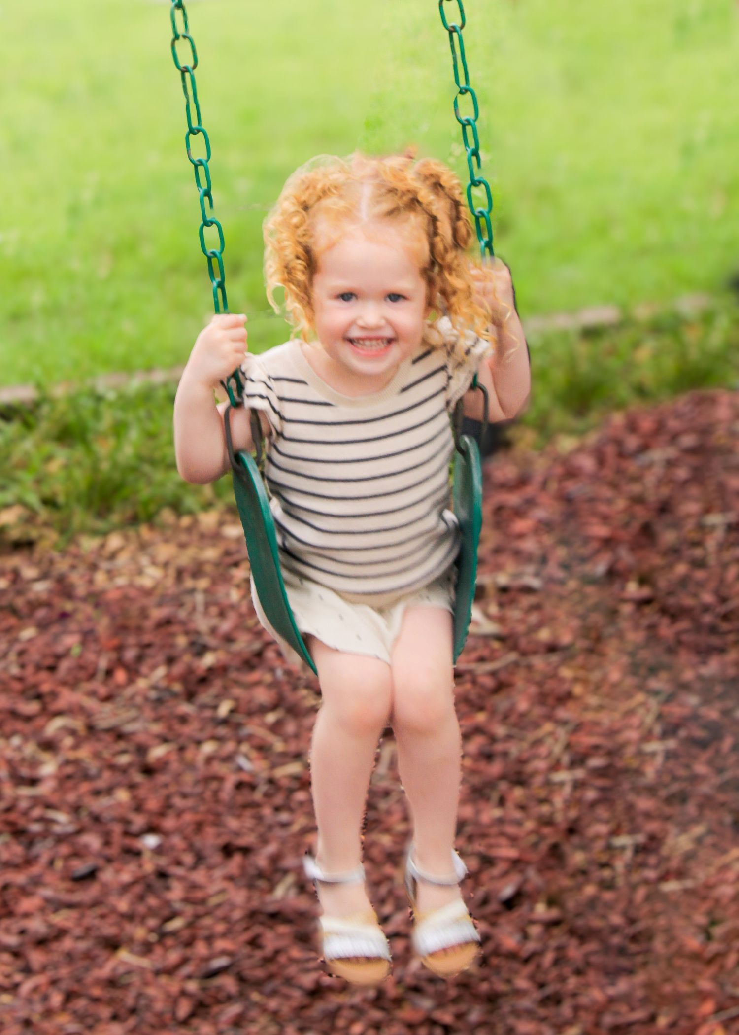 Child in striped outfit enjoying a green swing set against mulch ground covering.