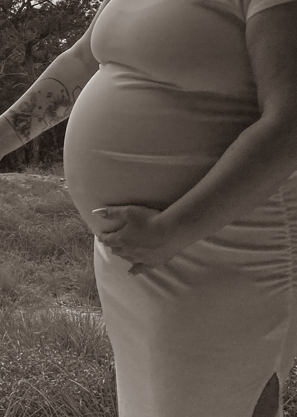 Black and white side profile of a pregnant belly with a tattooed arm resting on top.