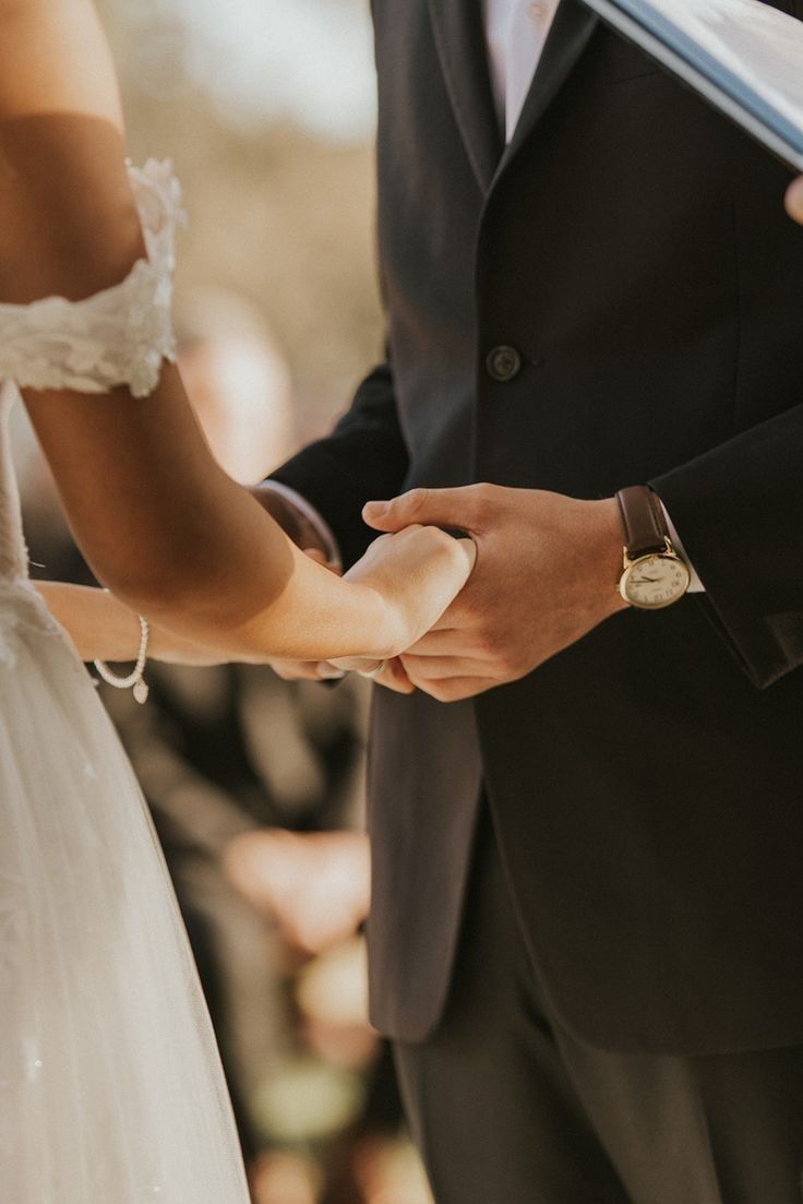 Close-up shot of bride and groom holding hands during wedding ceremony, with white lace dress and black suit visible.